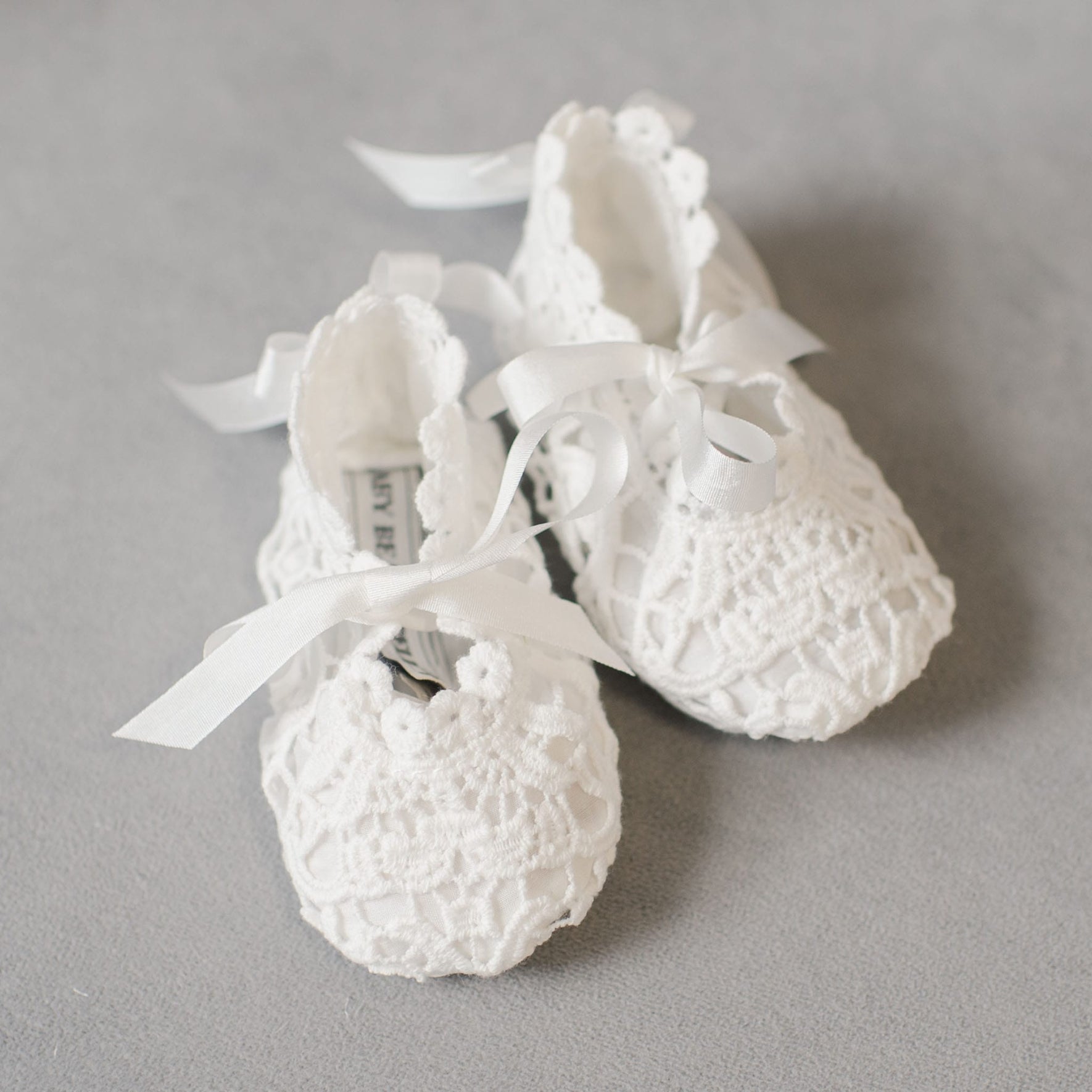 A pair of Grace Lace Christening Booties, featuring white crocheted fabric with ribbon ties, displayed on a gray surface.