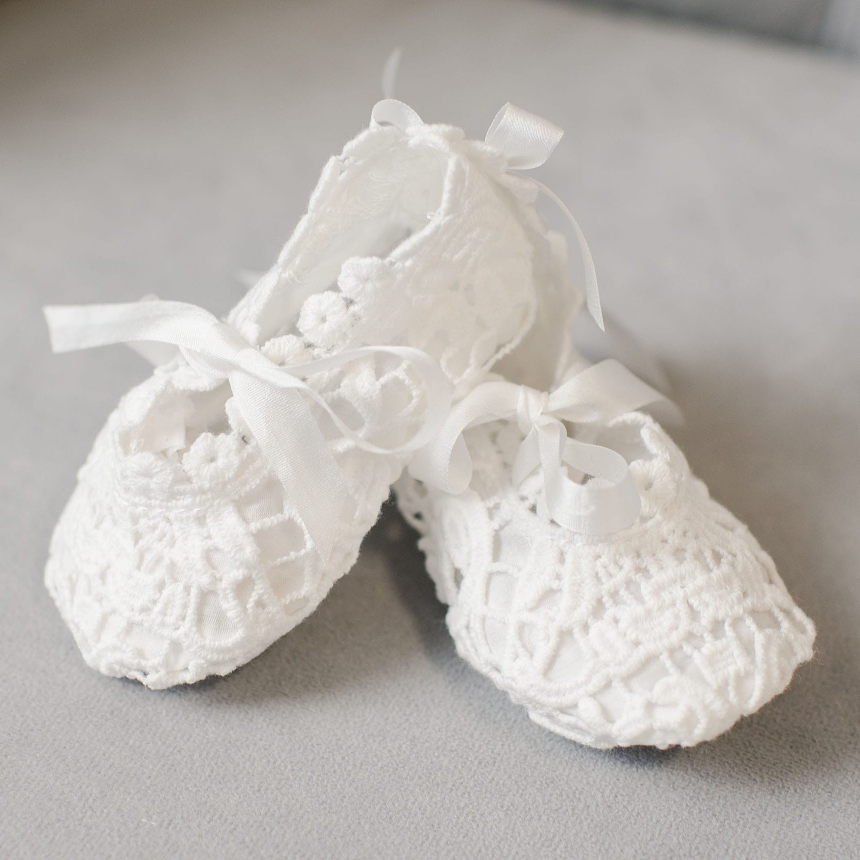 A pair of Grace Lace Christening Booties with ribbon ties are placed on a gray surface. These booties feature intricate patterns and a delicate design, perfect for completing a Christening Outfit.