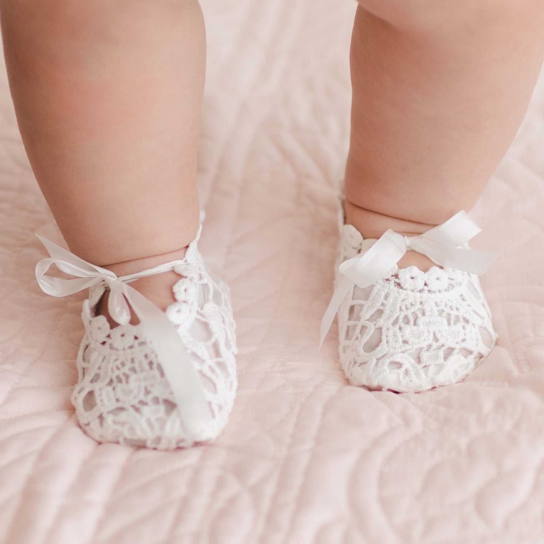 Close-up of a baby's feet wearing Grace Lace Christening Booties with ribbon ties on a soft pink quilted surface. The baby's chubby legs and the delicate details of the booties are clearly visible, ideal for a special occasion.
