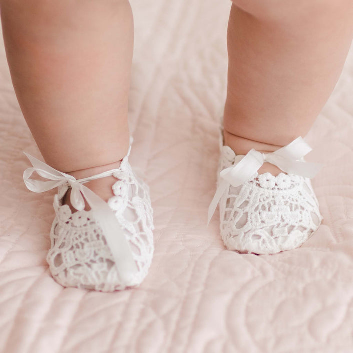 Close-up of a baby's feet wearing Grace Lace Christening Booties with ribbon ties on a soft pink quilted surface. The baby's chubby legs and the delicate details of the booties are clearly visible, ideal for a special occasion.