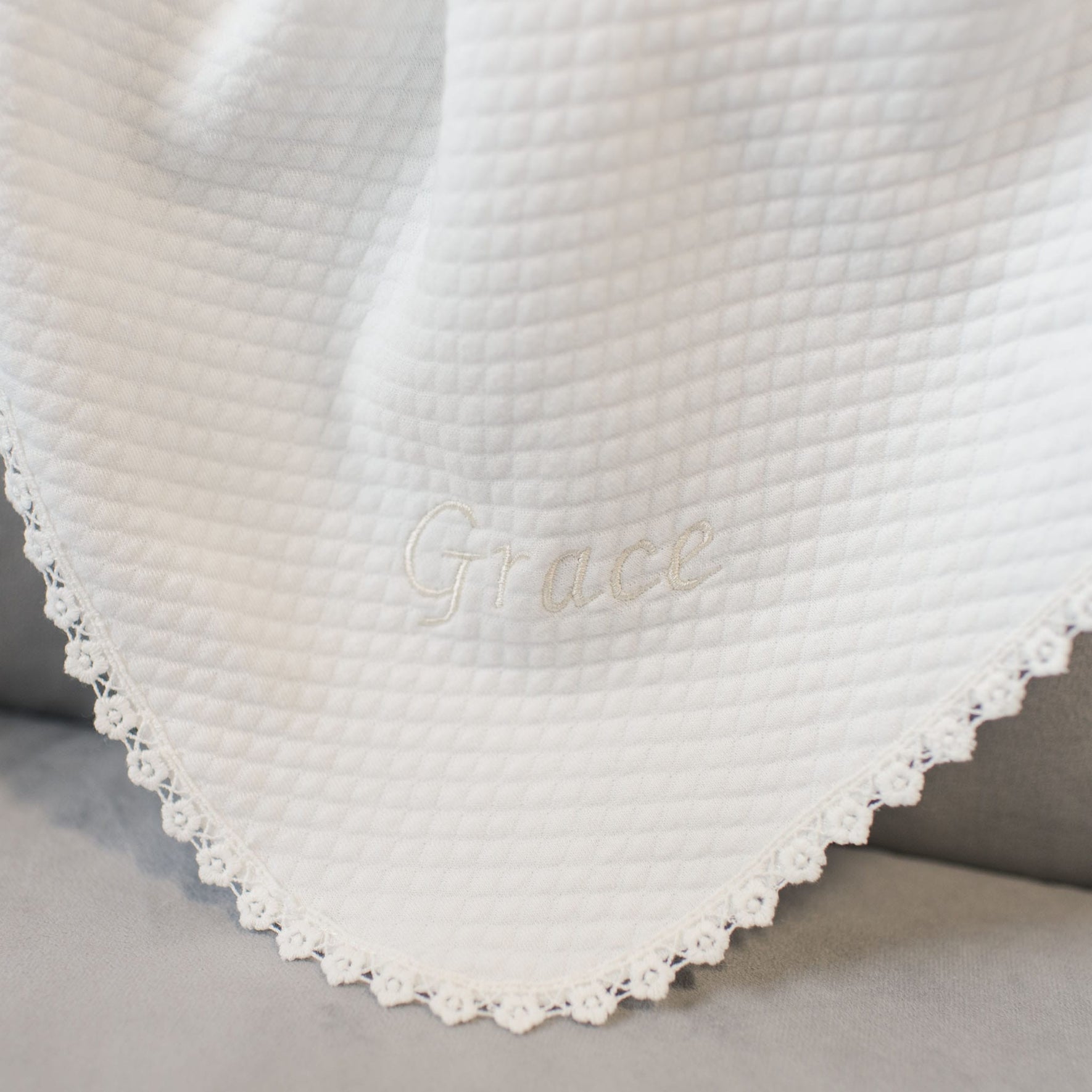 A Grace Personalized Blanket, soft and crafted from cotton with a white textured finish and scalloped lace trim, is draped over a surface. The name "Grace" is embroidered in the center of this handmade Christening blanket, designed to be a cherished heirloom piece.