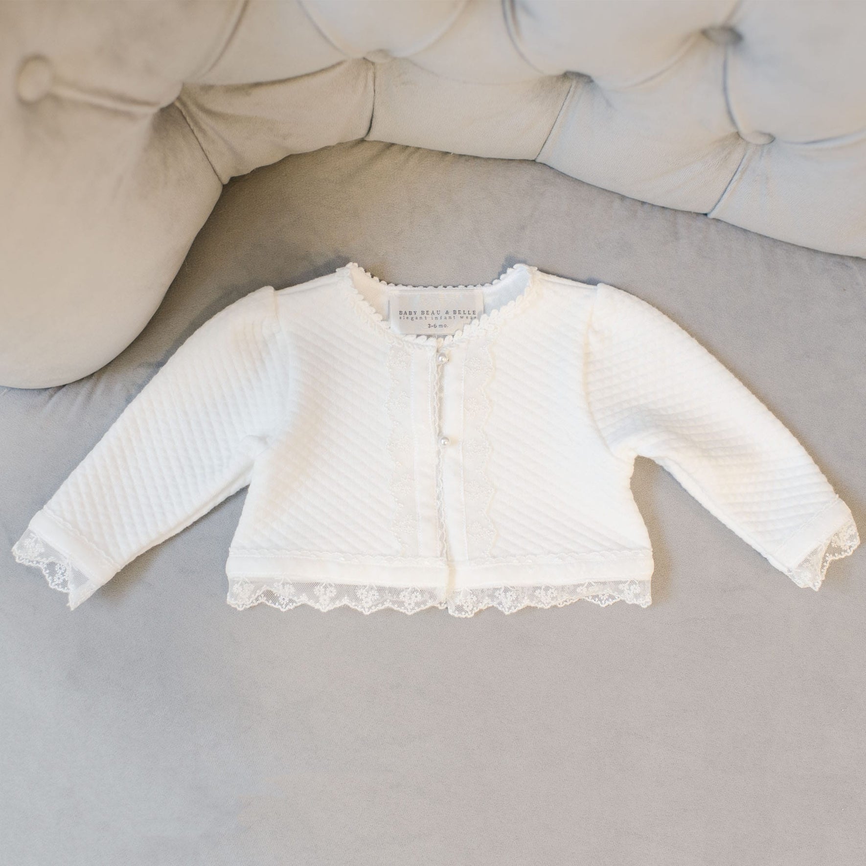 The Grace Quilted Cotton Sweater, adorned with floral lace trim and pearl-style buttons, is displayed on a soft, tufted gray surface. This white baby cardigan showcases a textured pattern.