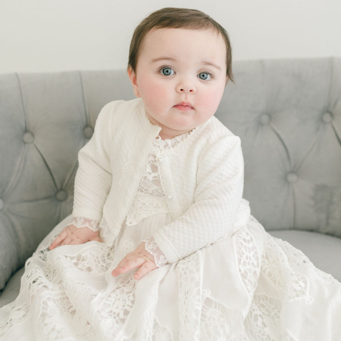 A baby wearing a white floral lace dress and the Grace Quilted Cotton Sweater with pearl style buttons sits on a light gray tufted couch. The baby has short dark hair and looks directly at the camera against a plain white background.