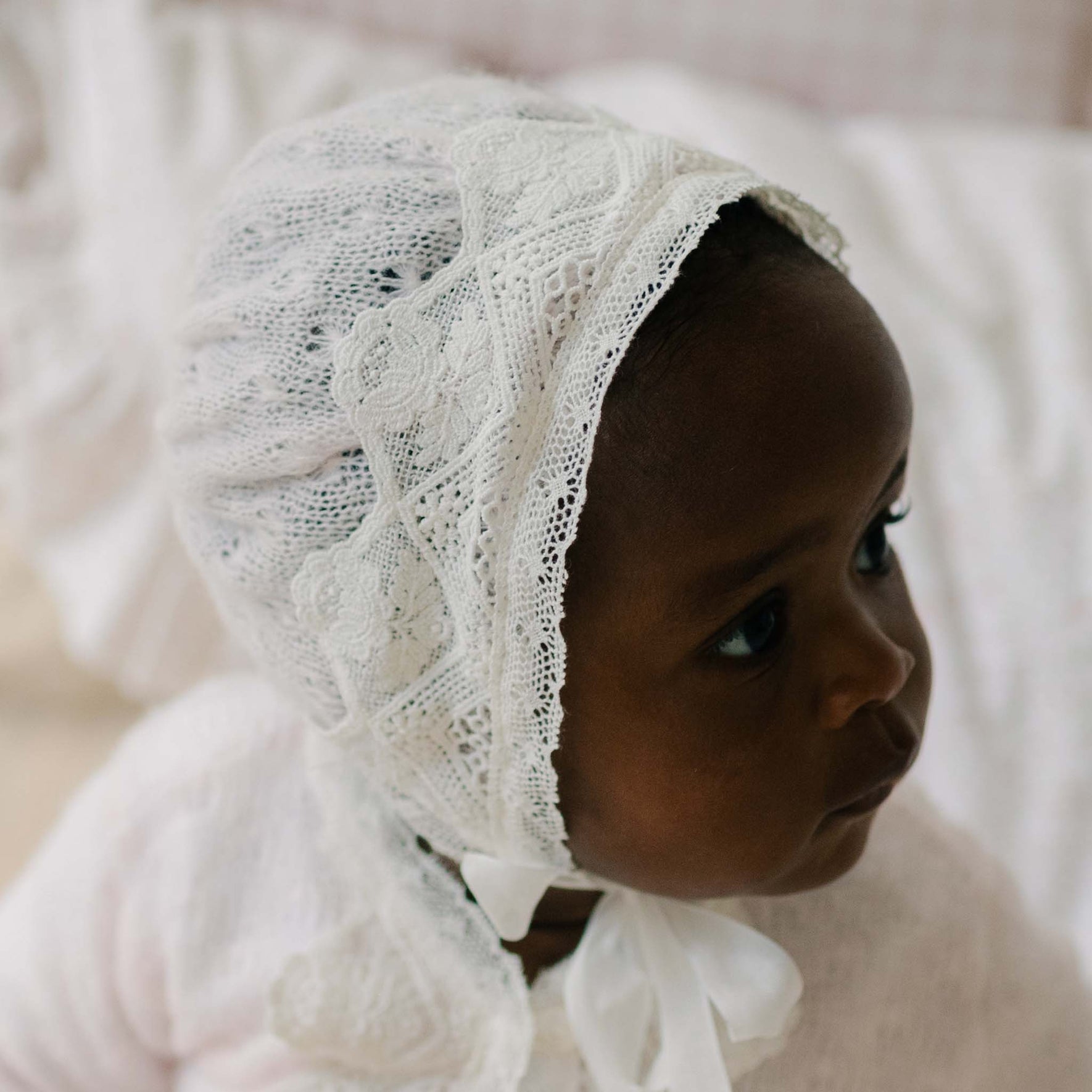 Close up of a baby wearing the Hailey Knit Bonnet against a soft, blurred background. Details show the knit and matching light ivory laces. 