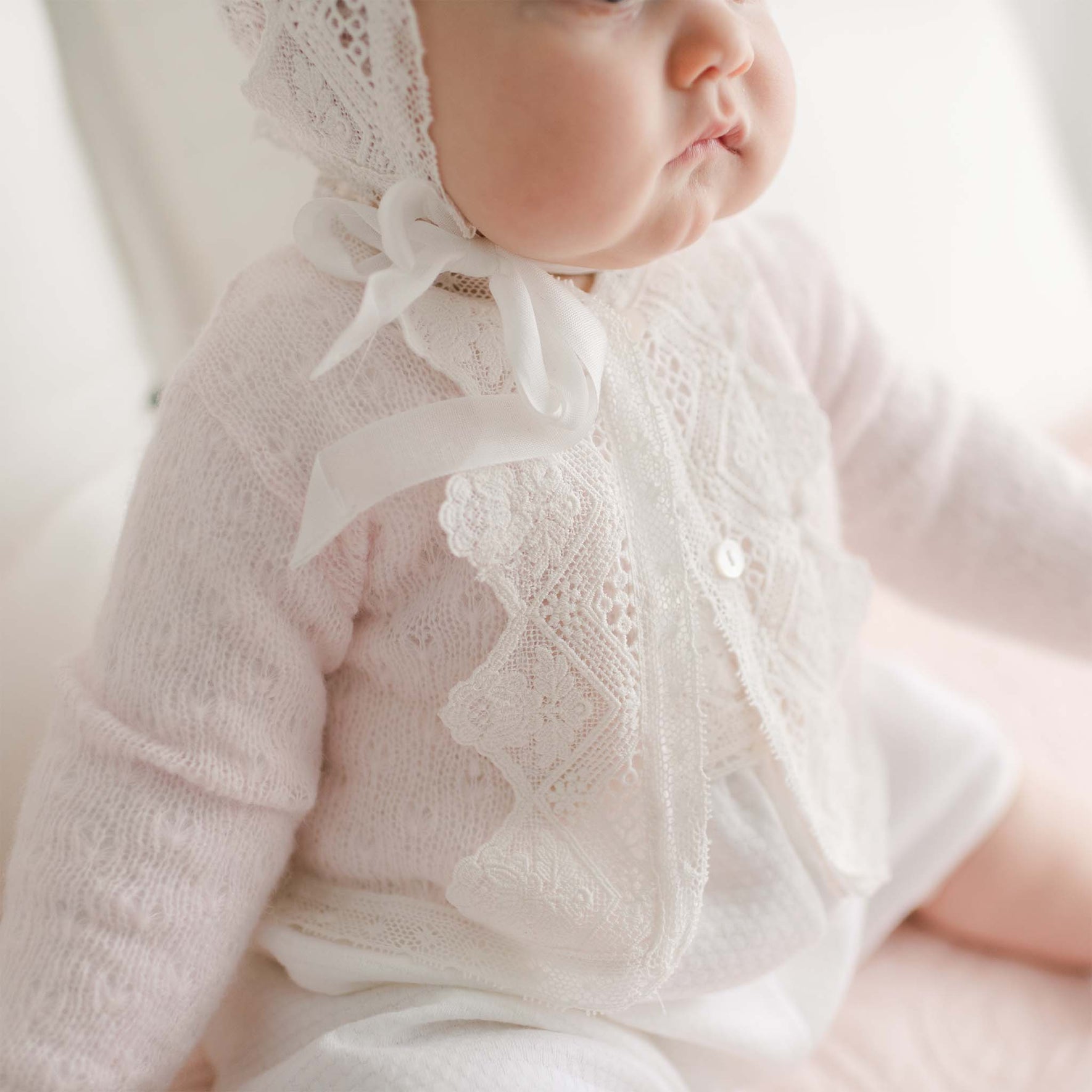 A baby dressed in a delicate, light pink Hailey Knit Sweater with white accents and a white bonnet sits with their head slightly turned. The atmosphere is soft and serene, highlighting the intricate knit acrylic details of the handmade USA baby's outfit.