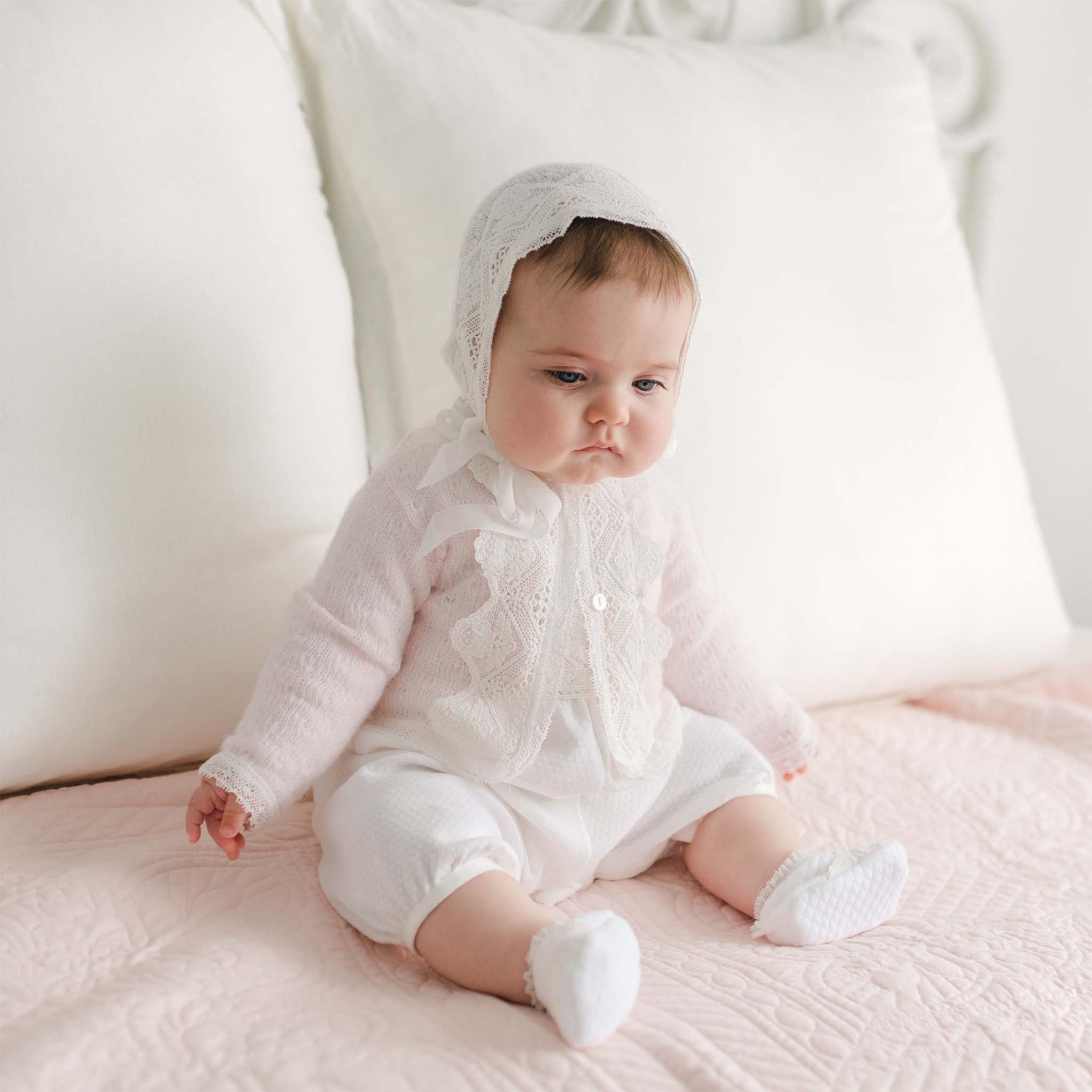 A baby dressed in a white Hailey Knit Sweater and bonnet sits on a bed with light pink covers and large white pillows in the background. The baby has a serious expression and is looking slightly to the right.