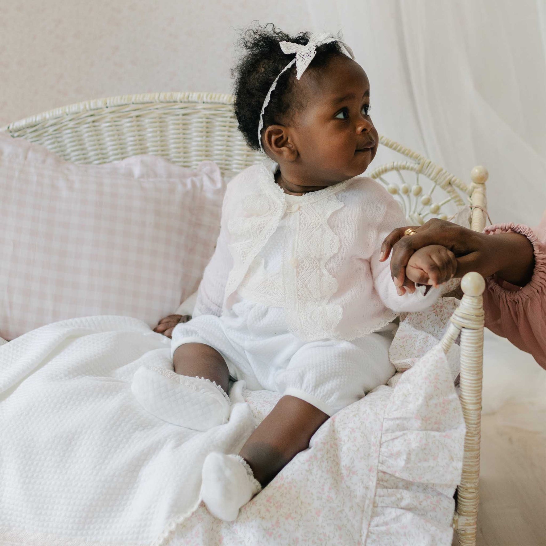 Baby in the Hailey Romper and matching Lace Headband sits in a white wicker chair with a pink patterned blanket. 