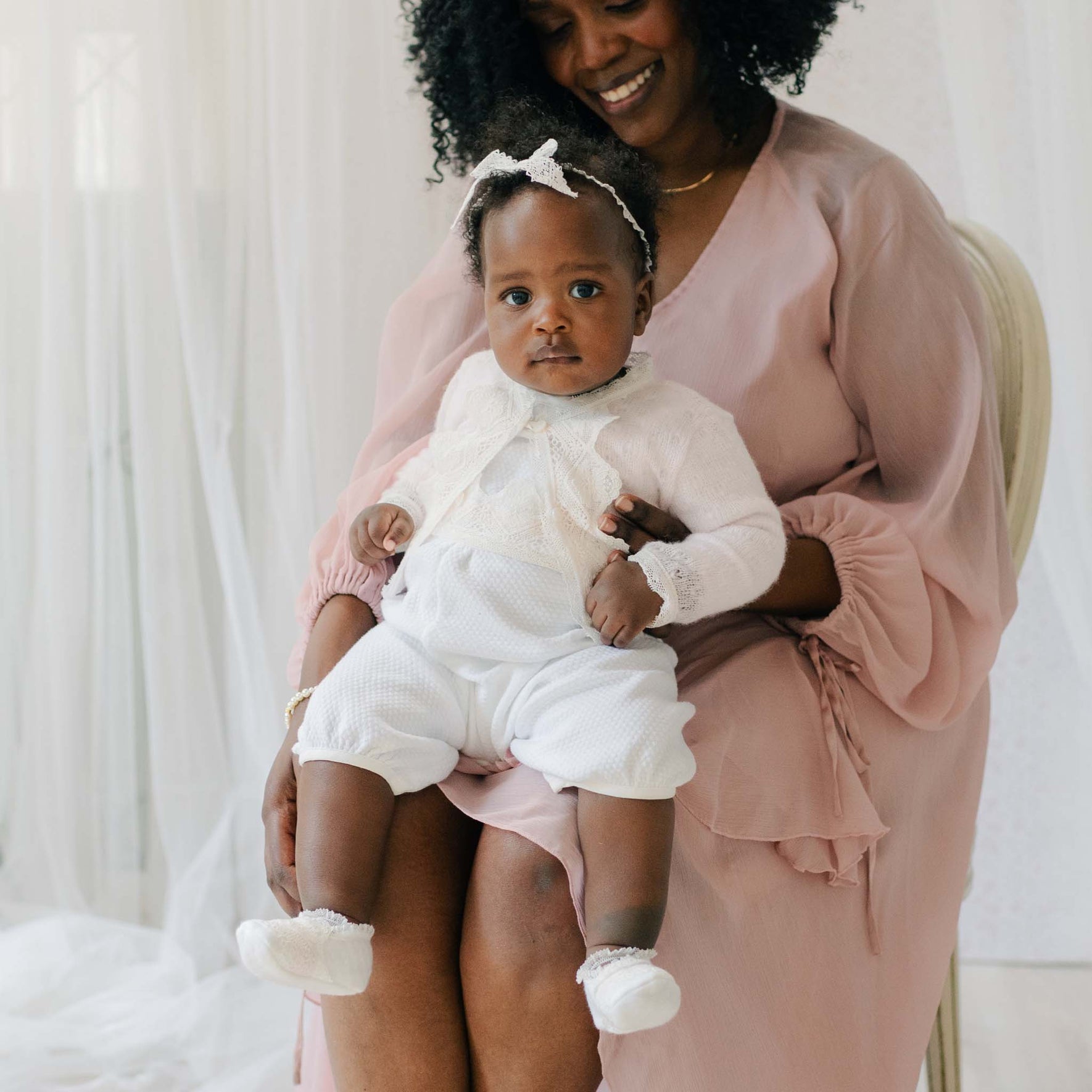 Baby wearing the Hailey Romper and matching Lace Headband. She is gently held by a woman in a pink dress against a white sheer background.