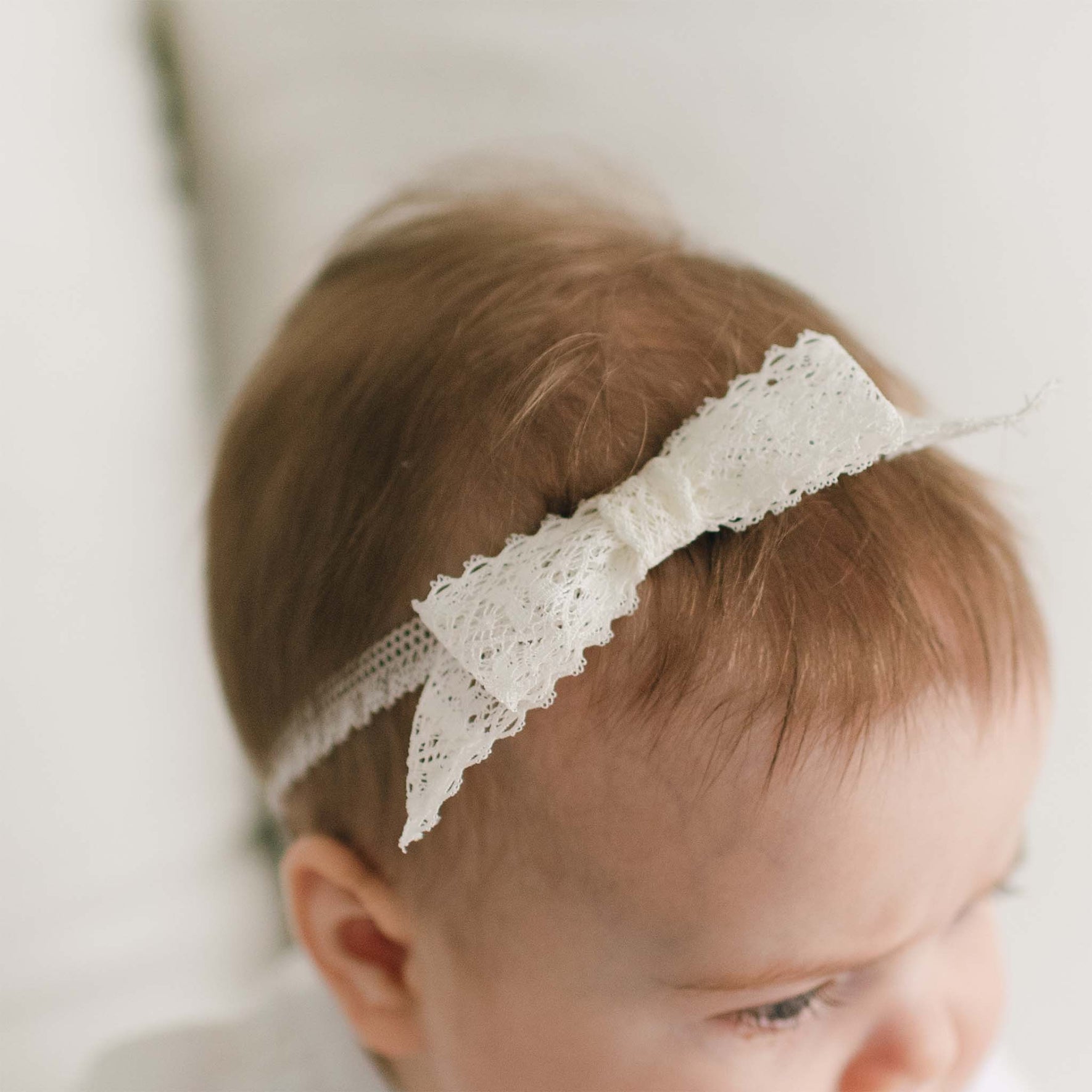 An infant with light brown hair is wearing a delicate Hailey Lace Headband tied in a bow. The baby is partially turned to the side, and their face is not fully visible. The background is a soft, neutral color.