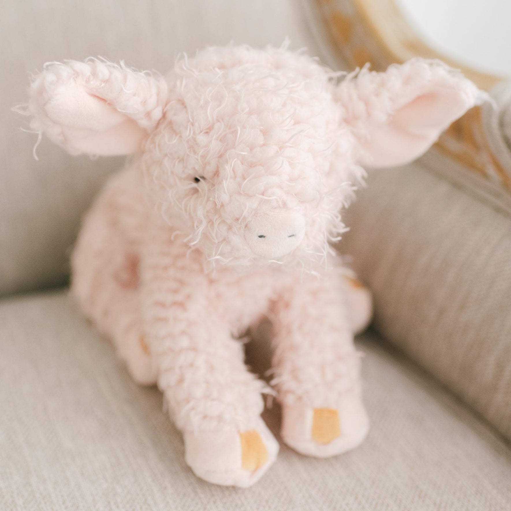 A plush toy pig named Hammie, featuring soft pink scraggle fur and an embroidered face, sits on a beige fabric chair. Its floppy ears and small black eyes enhance its cute appearance, making it a perfect baby gift. The background highlights a cushioned surface, adding to the cozy scene.