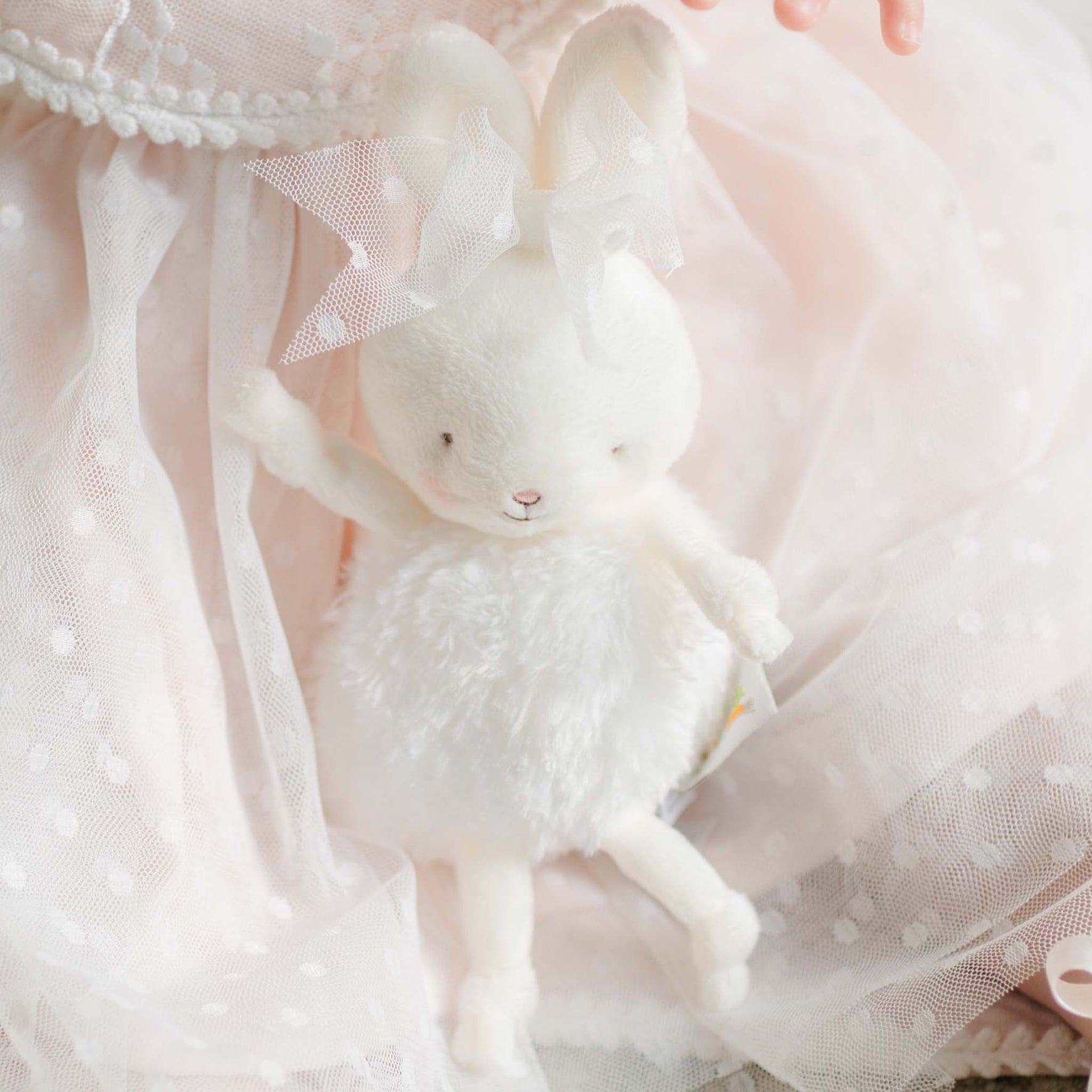 A plush white bunny with a fluffy body, soft embroidered features, and a delicate lace bow on its ear, resting against sheer dotted tulle fabric.