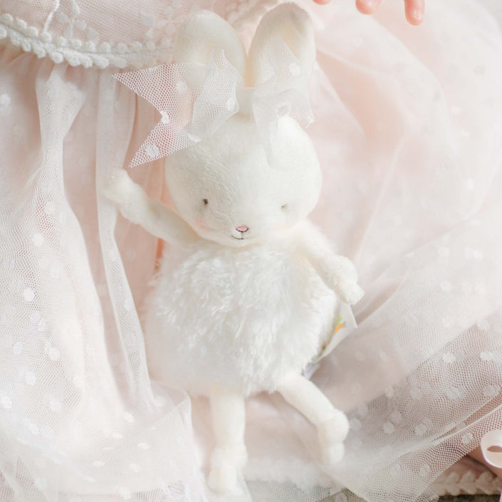 A plush white bunny with a fluffy body, soft embroidered features, and a delicate lace bow on its ear, resting against sheer dotted tulle fabric.