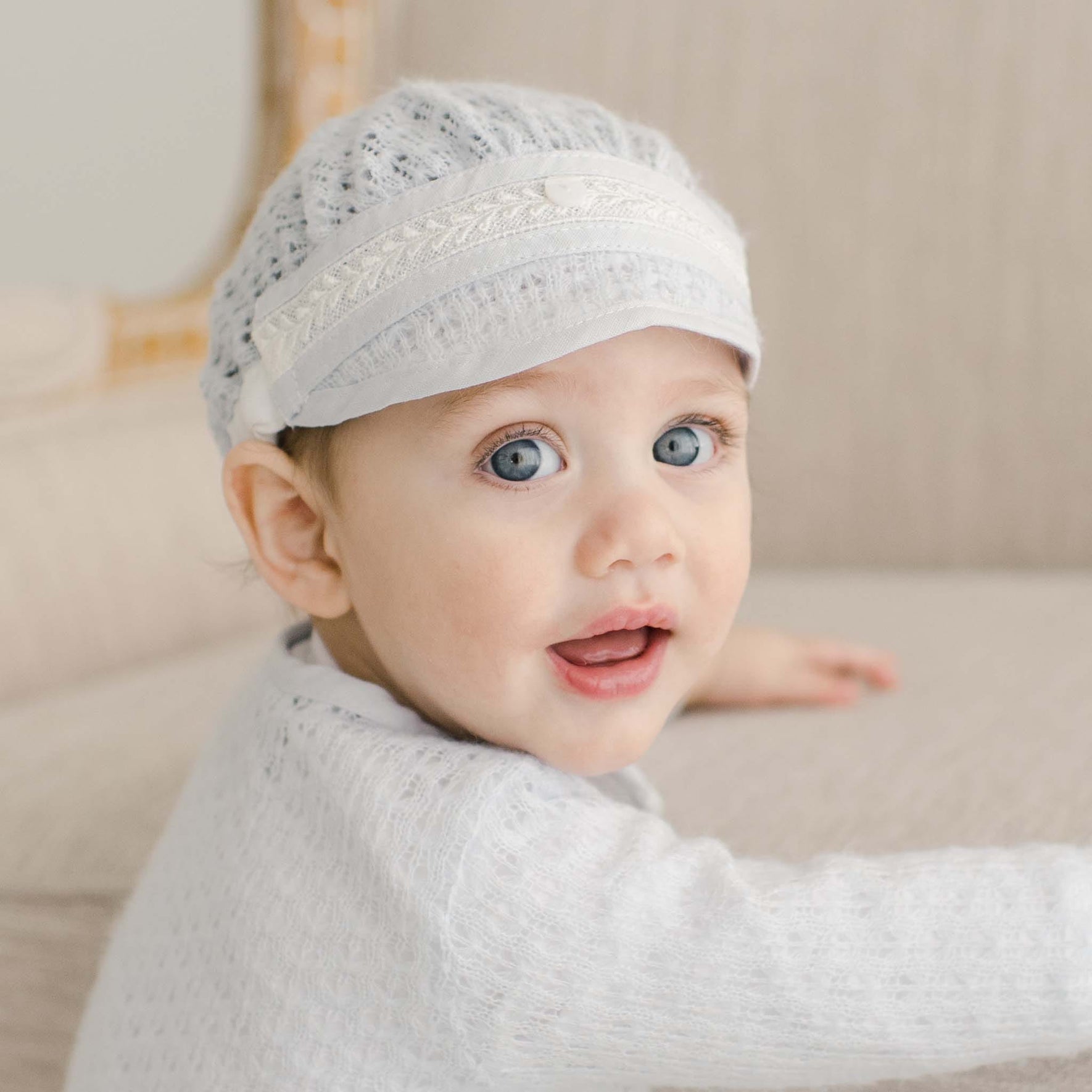 Baby in the Harrison Blue Knit Christening Hat, sitting on a beige chair, with exquisite lace details.
