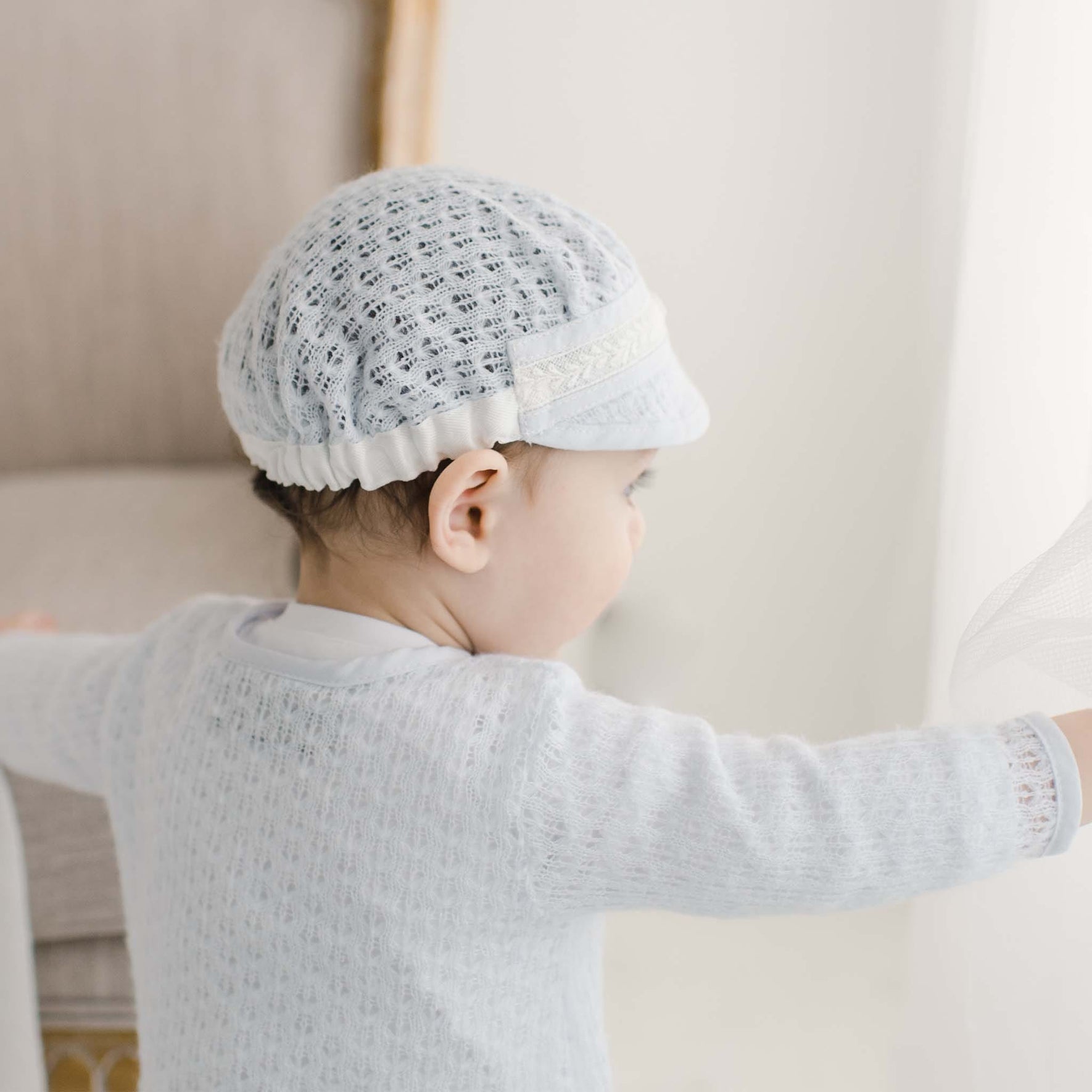  Back view of a baby dressed in a blue knit christening sweater and coordinating cap, highlighting the intricate lace accents and soft knit texture.