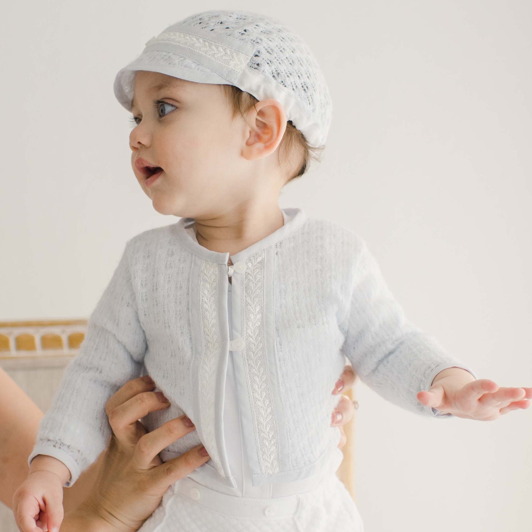 Baby boy in a blue knit christening sweater, held up to display the fine lace detailing and matching white attire.