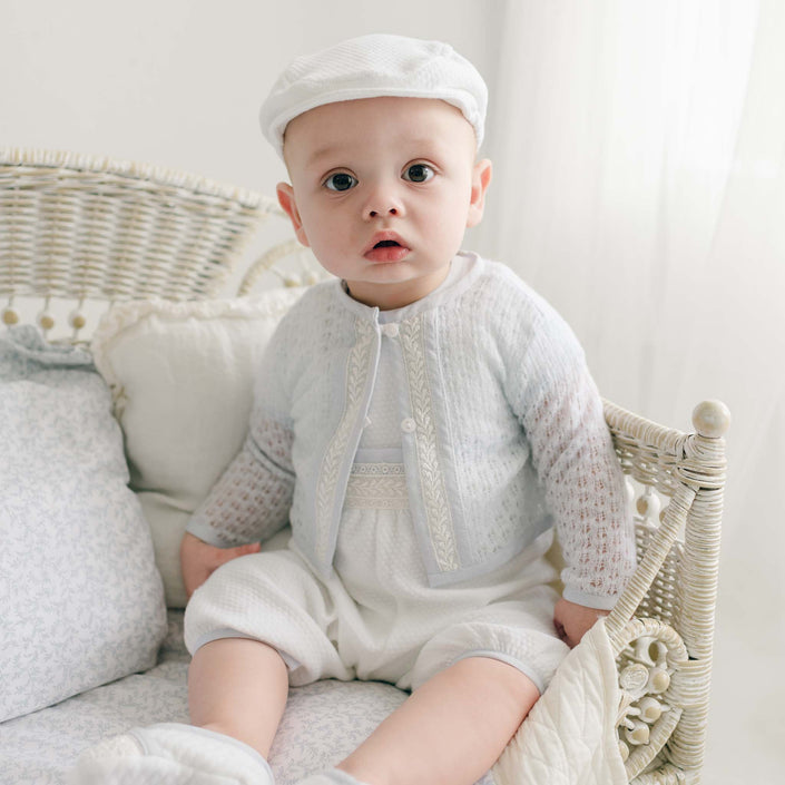 Baby boy in a wicker cradle wearing a blue knit christening sweater with lace trim and a white romper.