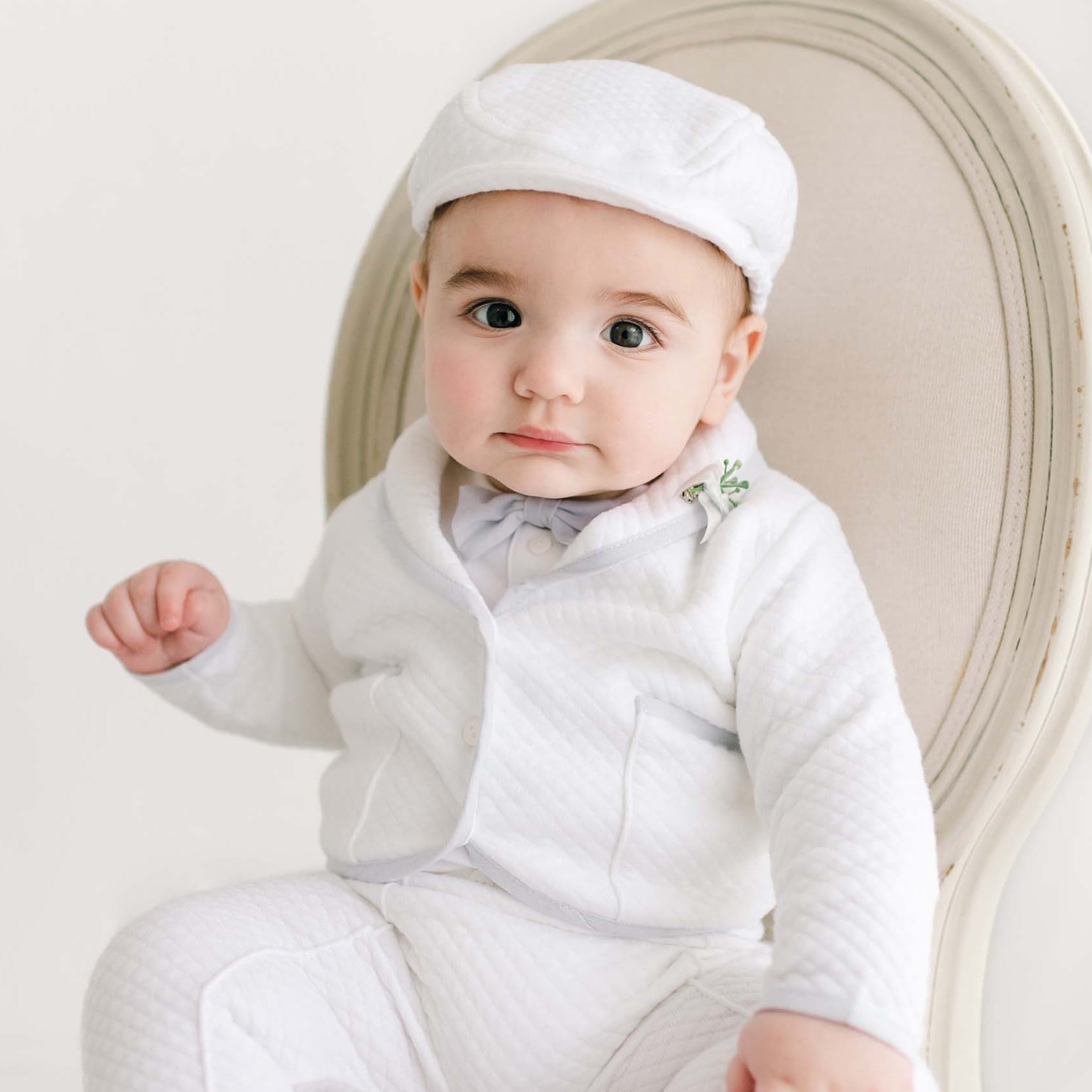 Infant wearing the Harrison Christening Pants Suit, styled with a white quilted jacket, pale blue trim, matching pants, a bow tie, and a classic cap.