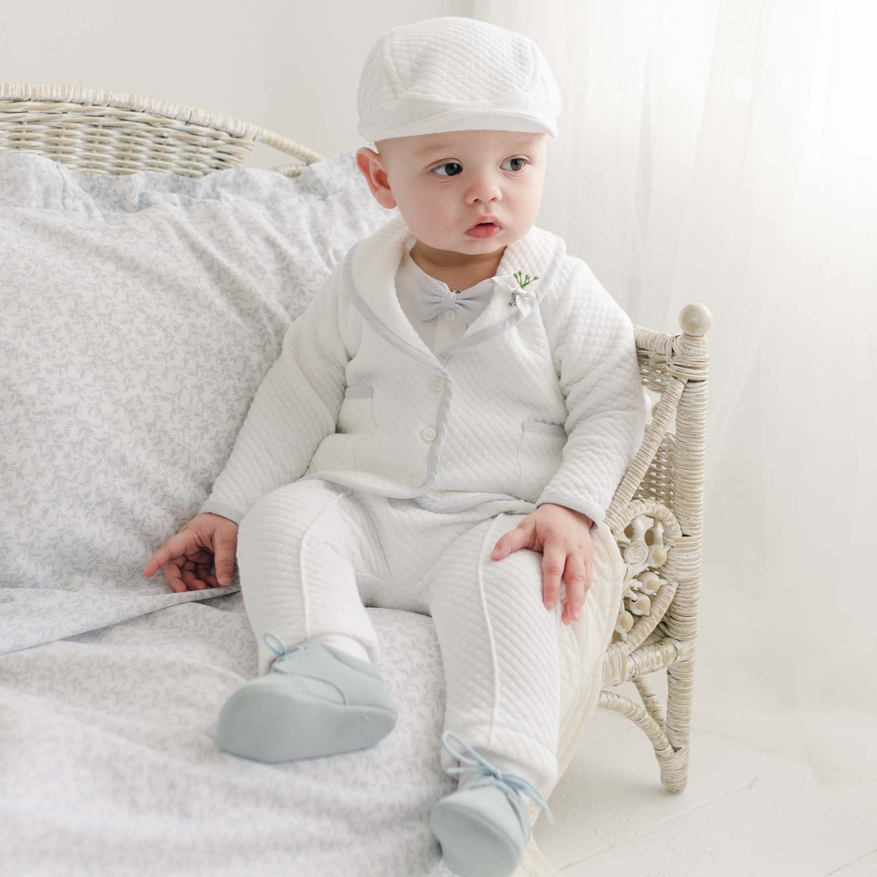 A baby boy dressed in the Harrison Christening Pants Suit is sitting on a light-colored wicker chair with cushioned bedding.