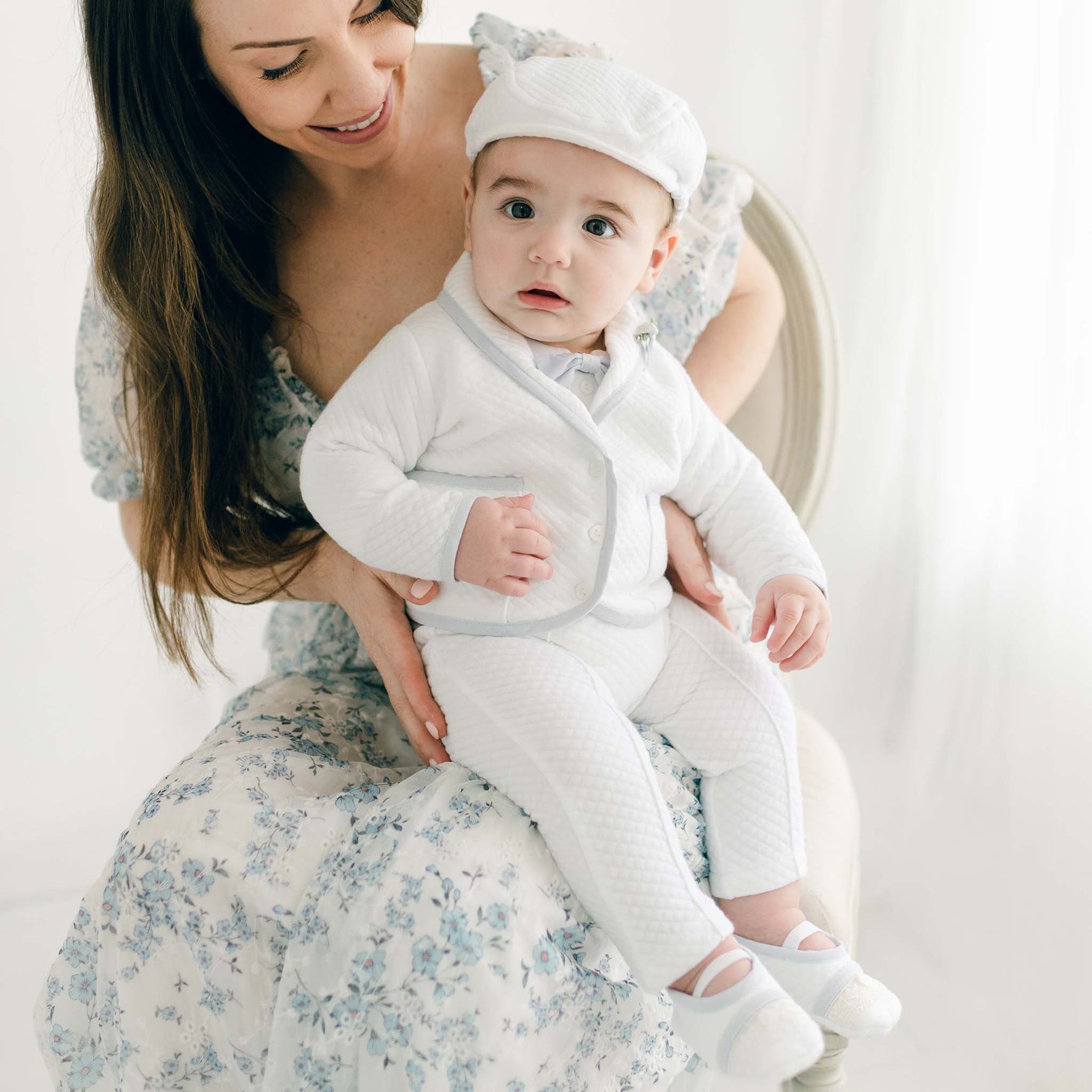 A mother in a floral dress holds her baby boy who is wearing the Harrison Christening Pants Suit. The baby sits on her lap, looking at the camera. 