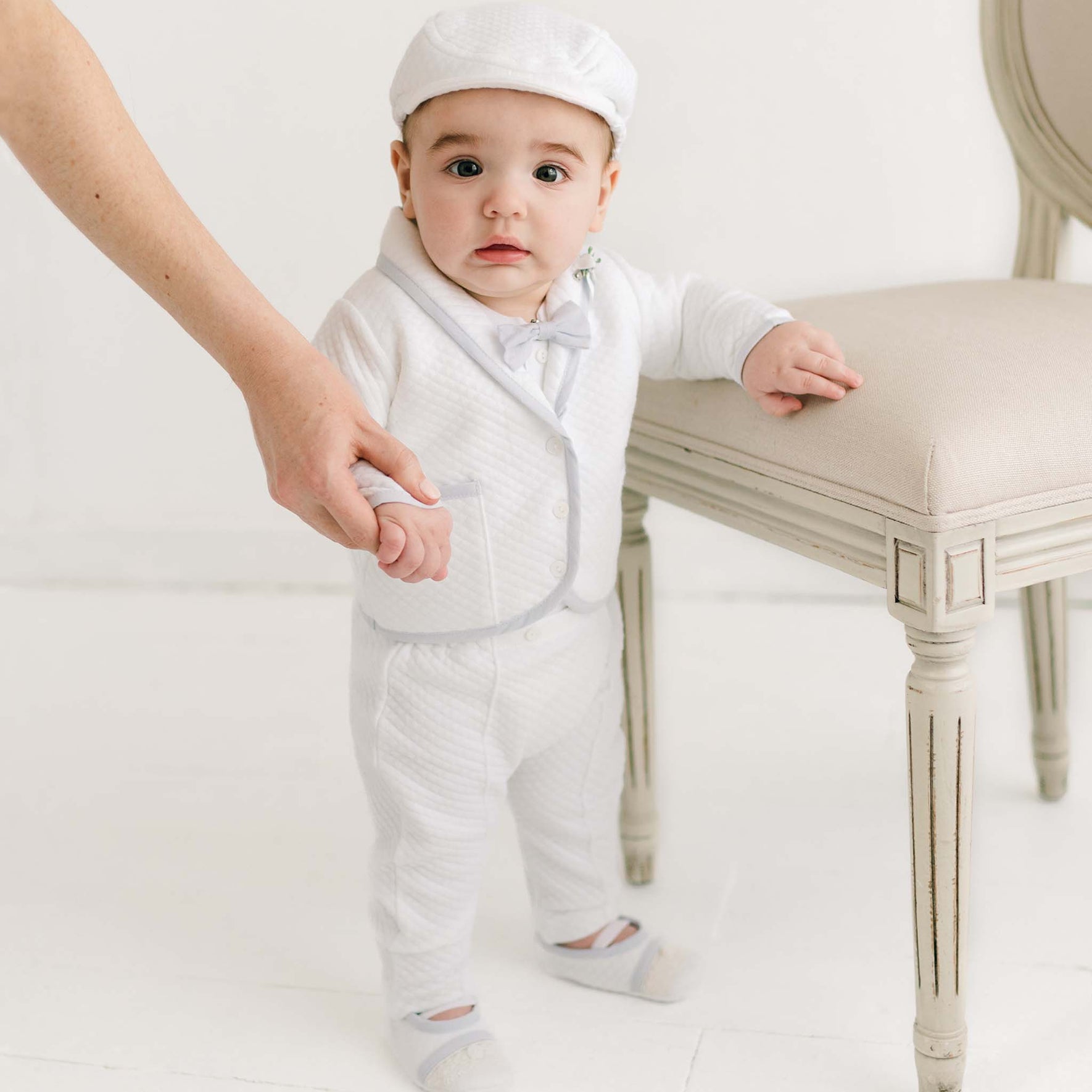 A baby, dressed in the Harrison Christening Pants Suit with the matching Newsboy Cap, is standing while holding onto an adult's hand.