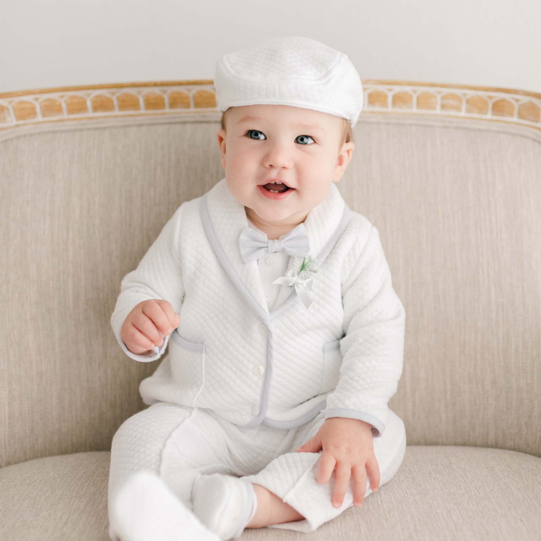 Baby boy seated on a beige sofa, wearing the Harrison Christening Pants Suit, a coordinating bowtie, and a small boutonniere detail.
