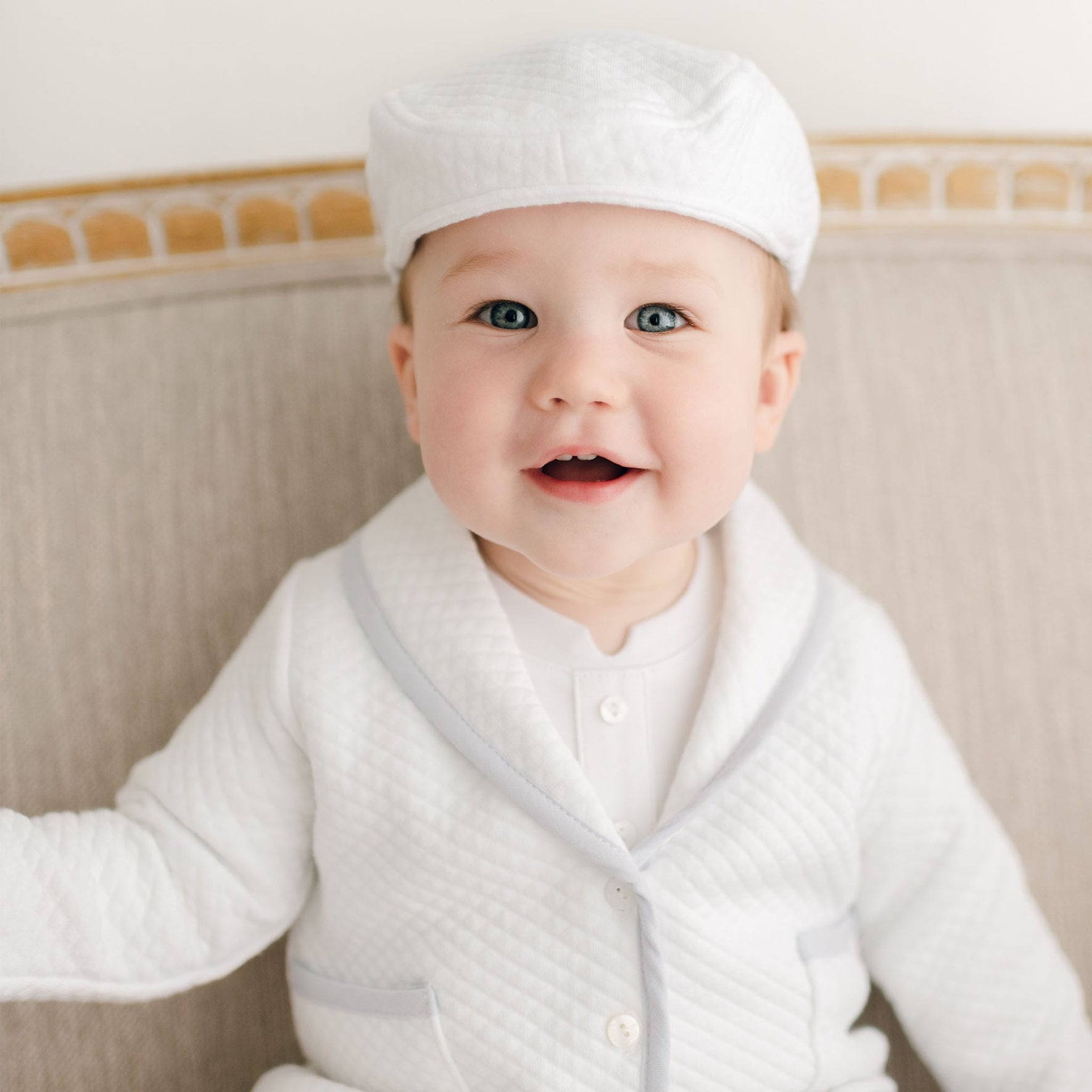 Smiling baby boy in the Harrison Christening Pants Suit, styled with a quilted white jacket, light blue trim, and matching cap, seated on a light beige sofa.