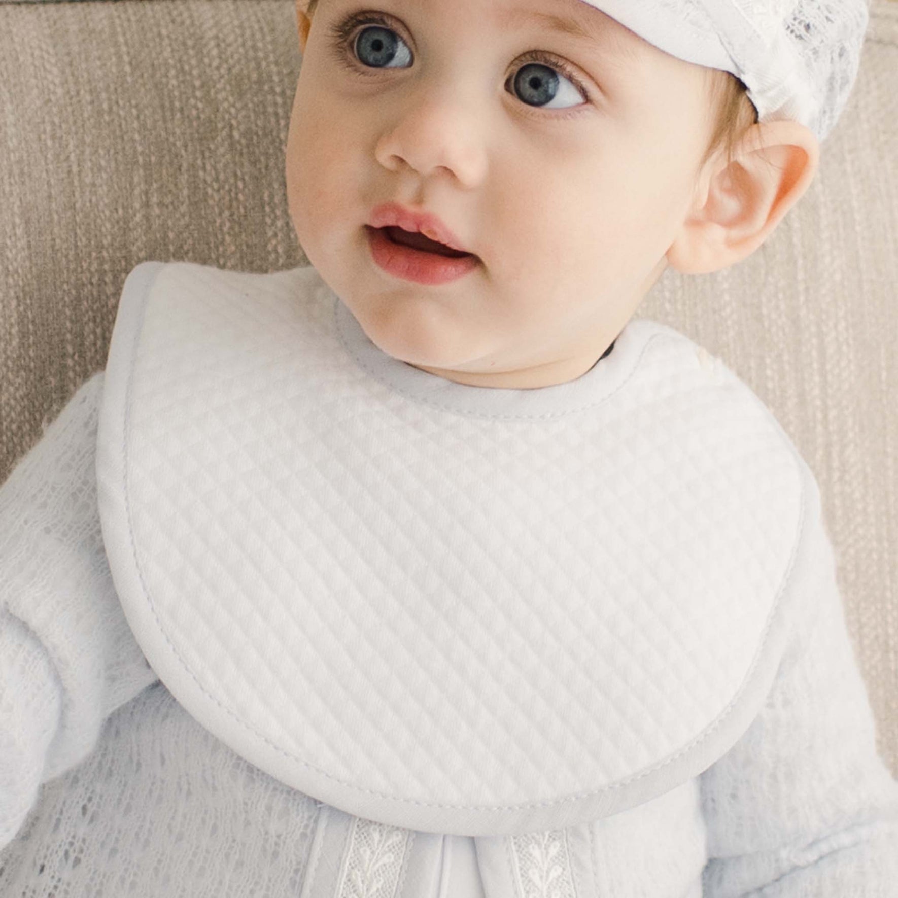 A baby wearing the Harrison Christening Bib, made of quilted white fabric with a blue trim.