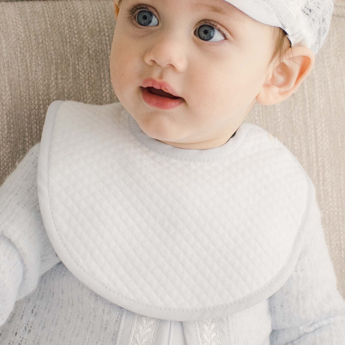 A baby wearing the Harrison Christening Bib, made of quilted white fabric with a blue trim.
