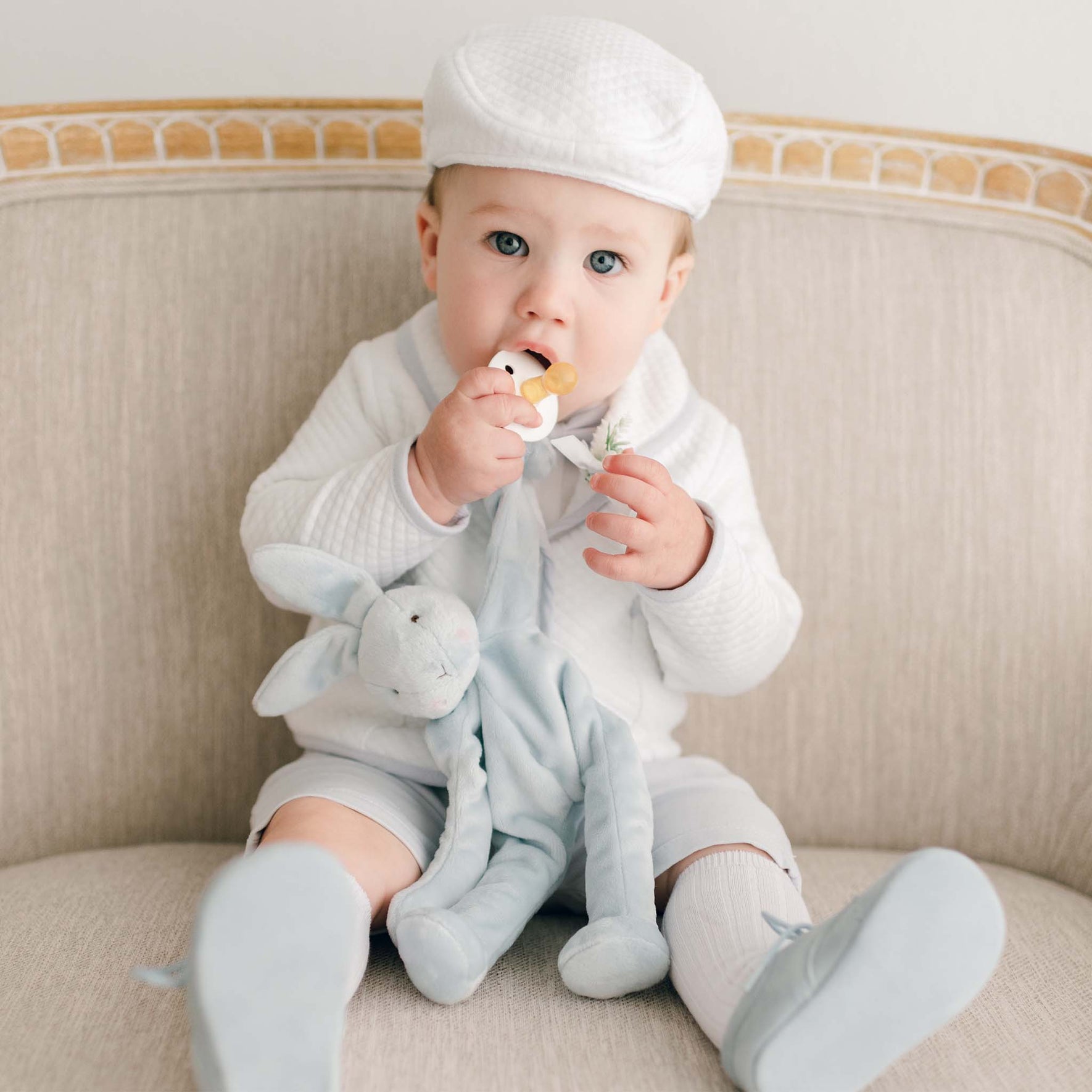 Baby boy dressed in the Harrison Christening Shorts Suit, holding a light blue bunny toy while styled with a quilted jacket, matching cap, and suede shoes.