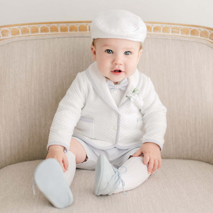 Baby boy dressed in the Harrison Christening Shorts Suit, featuring a white quilted jacket, matching shorts, and newsboy cap, with knee-high white socks and blue suede shoes.
