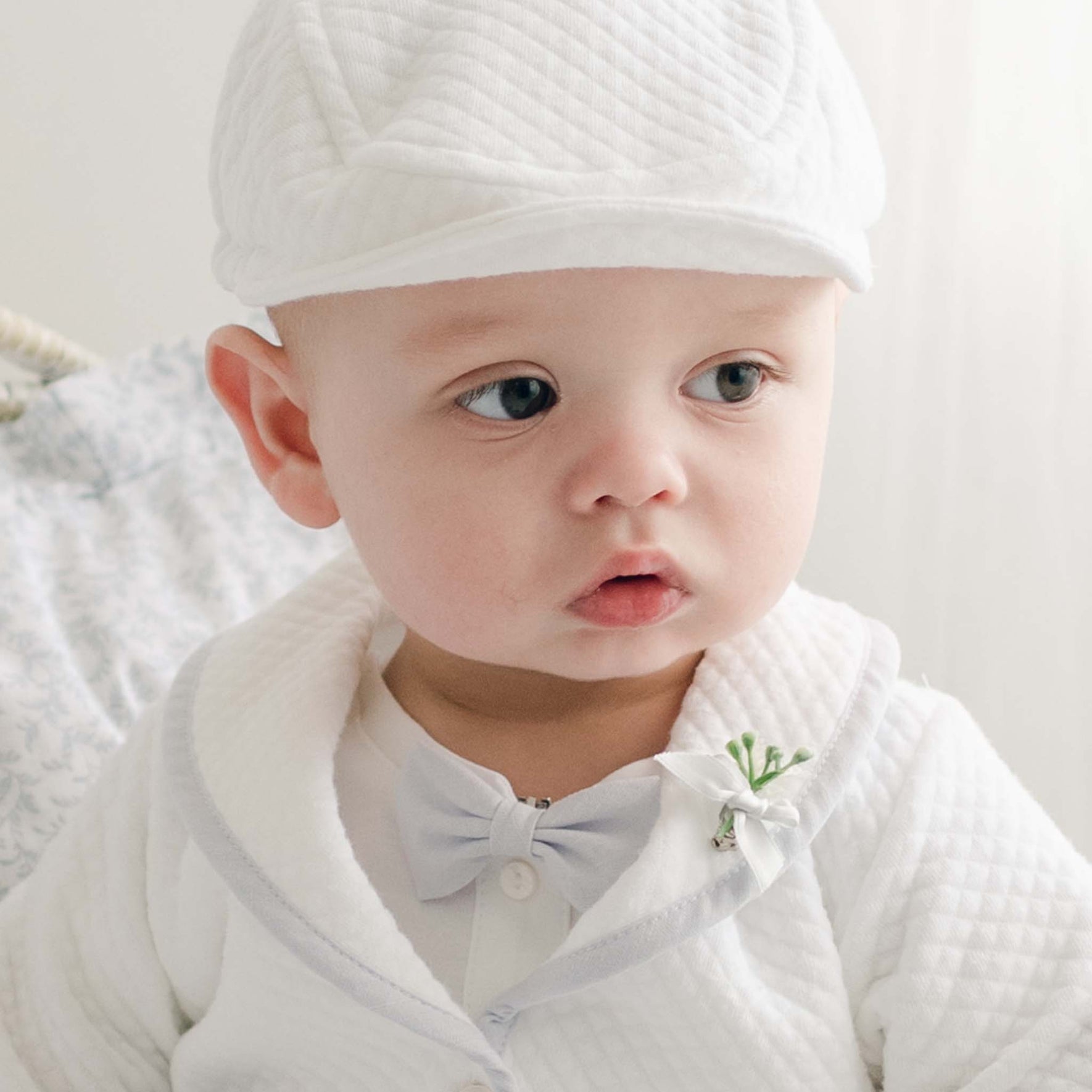 A baby boy wearing the Harrison Christening Bow Tie & Boutonniere with a quilted jacket and matching cap, perfect for a christening day ensemble.