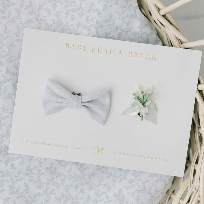 The Harrison Christening Bow Tie & Boutonniere set, showcasing a pale blue linen bow tie and a boutonniere with white ribbon and green sprigs, elegantly presented on a branded Baby Beau & Belle card.
