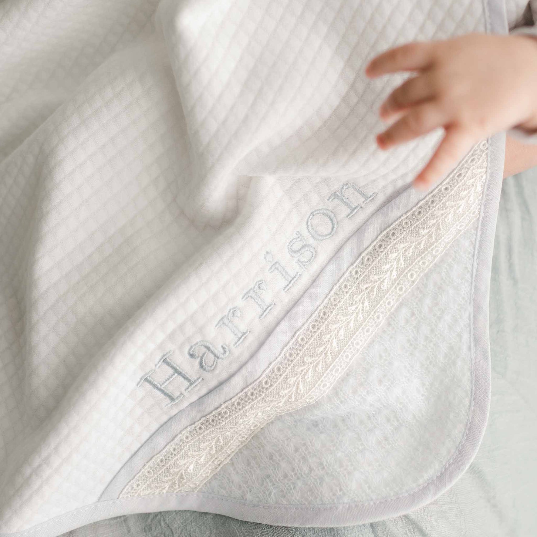 A baby's hand rests on a personalized quilted blanket featuring the embroidered name "Harrison" and elegant lace trim, beautifully complementing the Harrison Newborn Accessory Bundle - Save 15%.