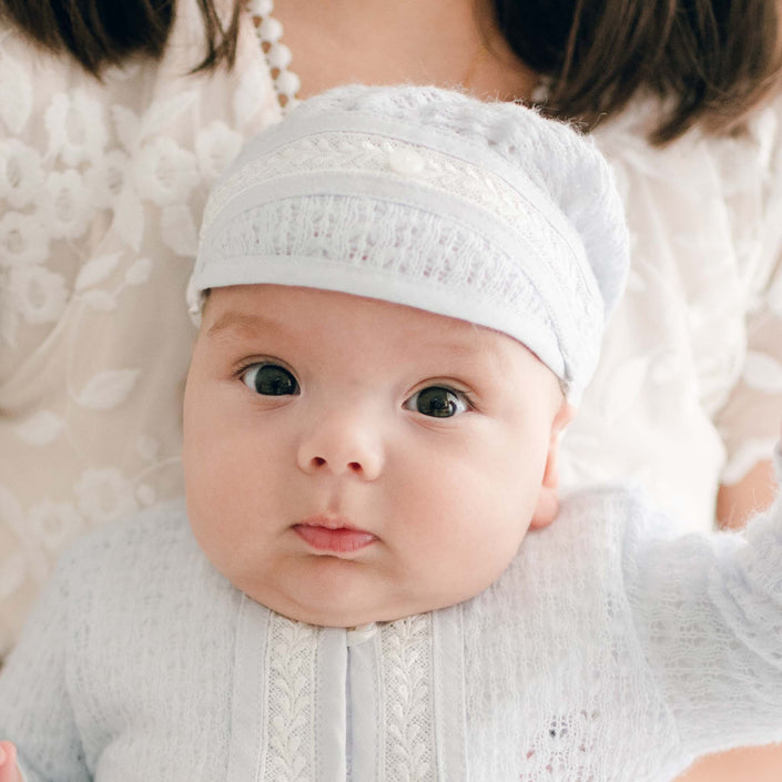 A baby wearing the Harrison Newborn Knit Hat and a light blue knitted outfit, reminiscent of a charming newborn baptism attire, is being held. The baby has wide eyes and chubby cheeks. The background is softly out of focus, featuring a person in a white outfit.