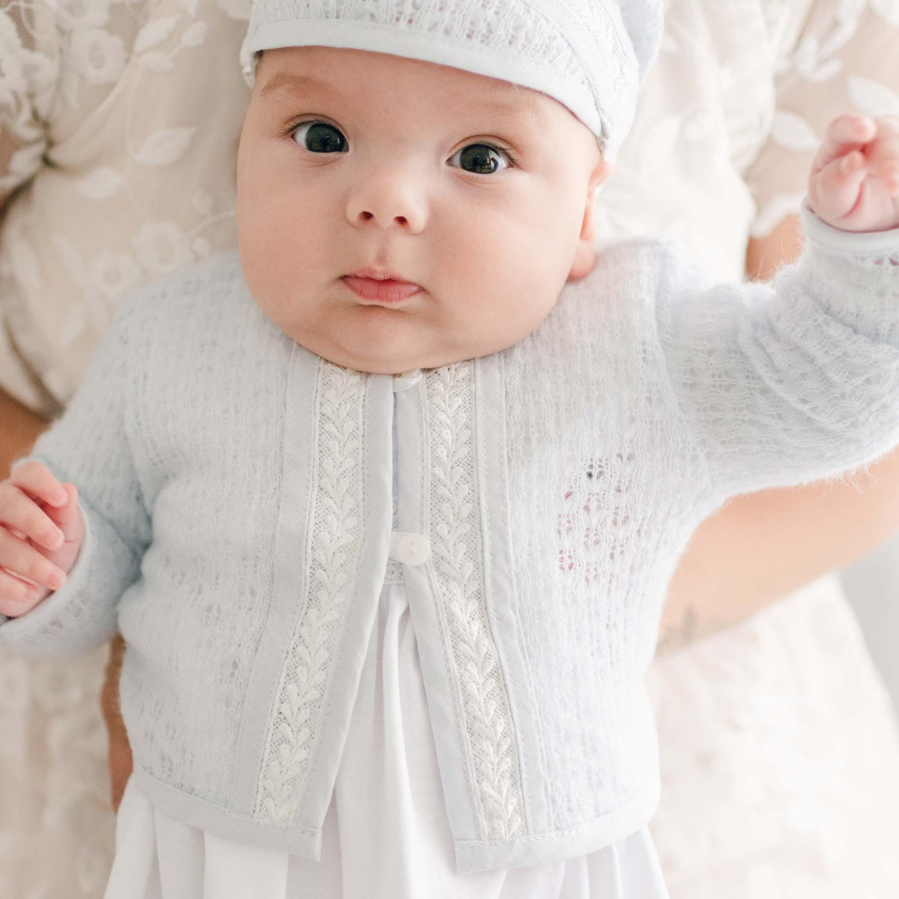 A baby wearing a Harrison Newborn Sweater in soft blue, complete with a matching hat, is gently held by an adult. The baby's wide eyes and slightly open mouth contrast with the softly blurred floral background.