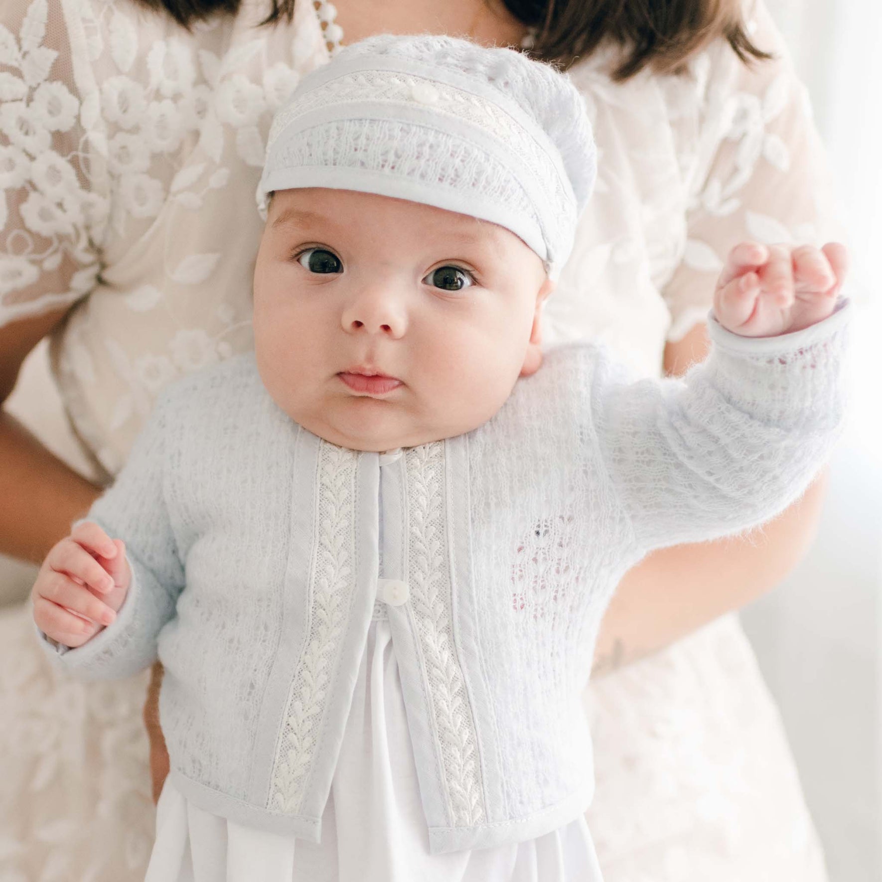 A baby, dressed in the Harrison Newborn Sweater—a soft blue knit baptism outfit with a matching hat—is held by an adult. The infant gazes directly at the camera, one arm raised excitedly. The adult is wearing a white lace garment against a softly lit background.