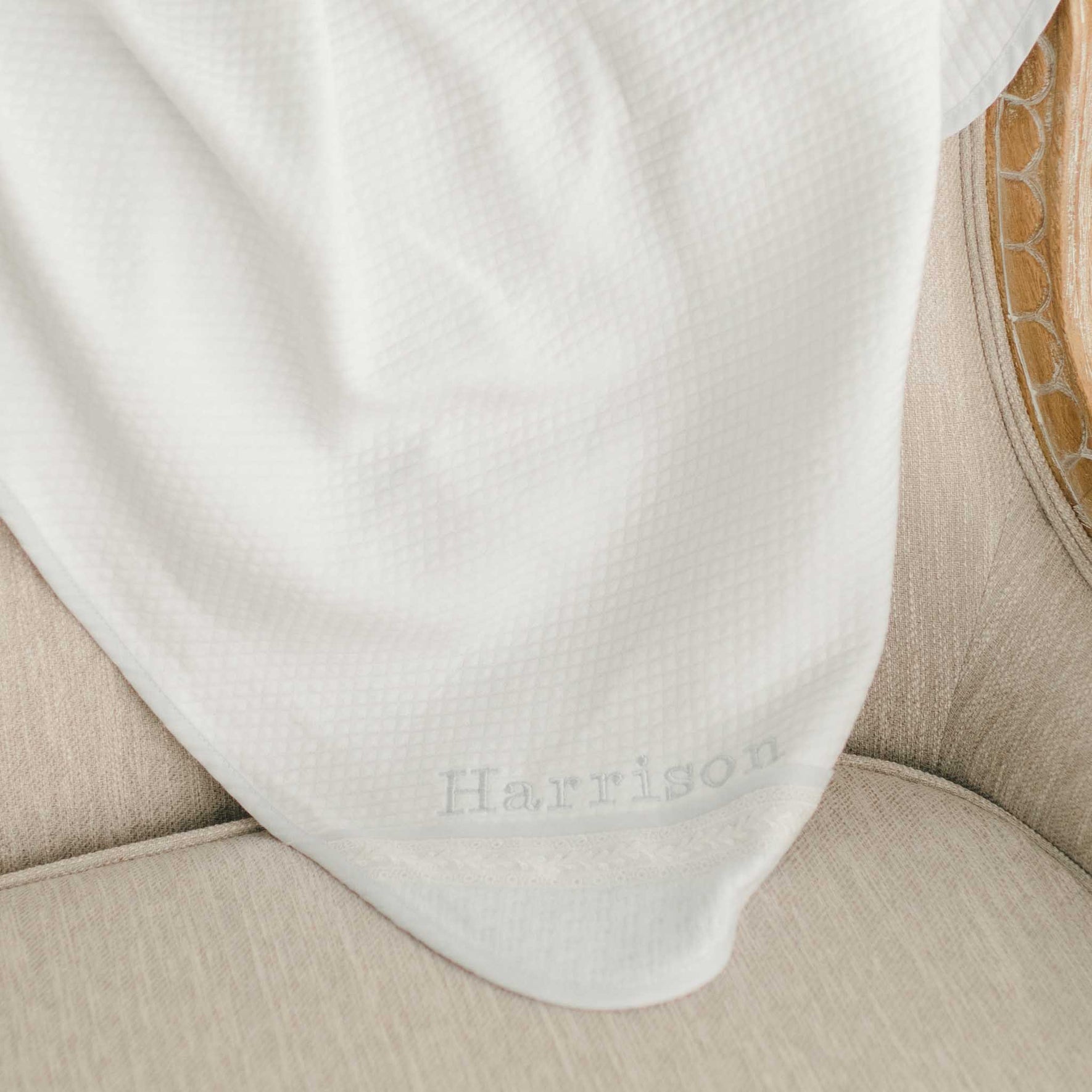A white quilted christening blanket, personalized with "Harrison" in light blue embroidery and featuring intricate lace edging, displayed draped over a chair.