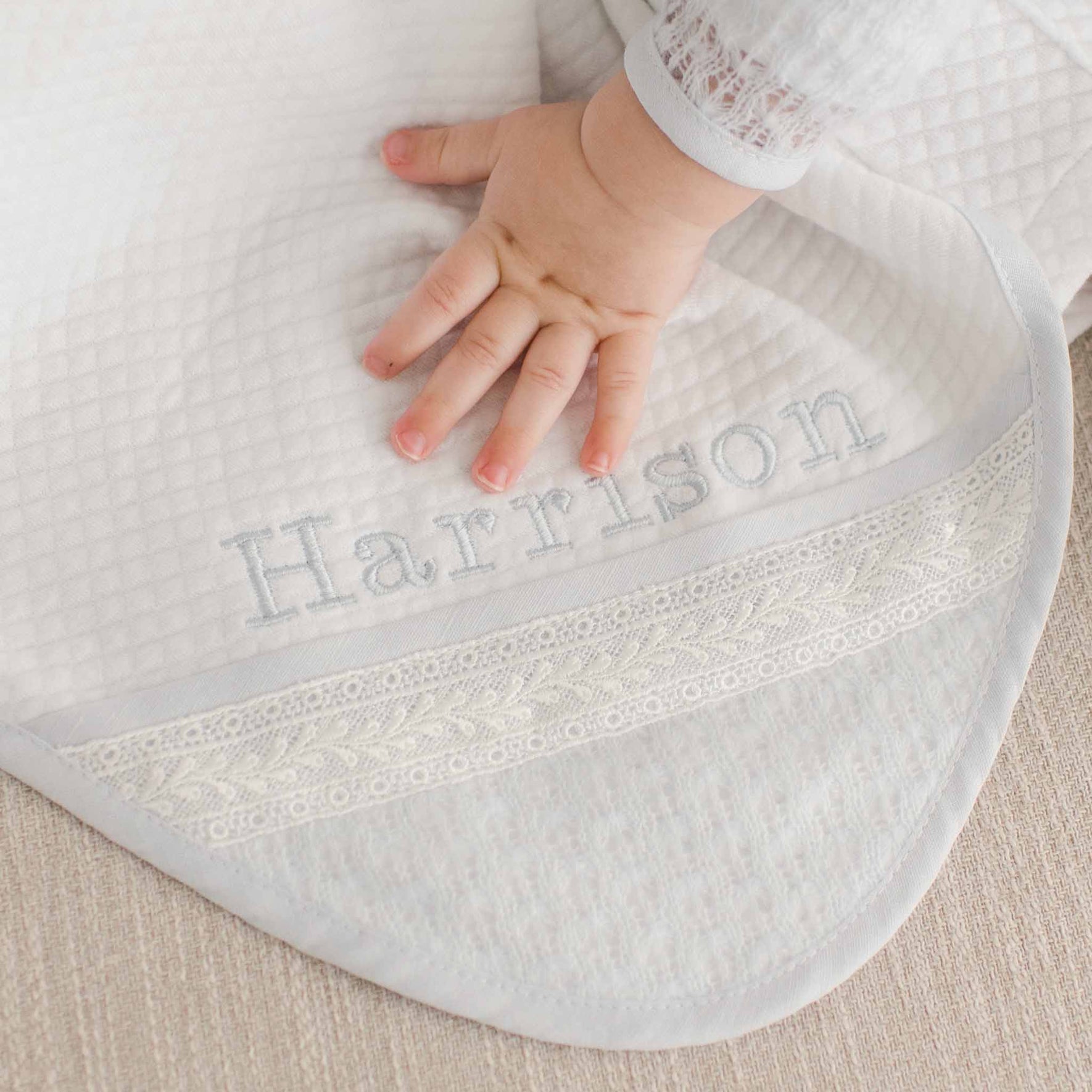 A folded white quilted blanket with "Harrison" embroidered in soft blue, accented by elegant lace trim, draped over a neutral-colored chair.