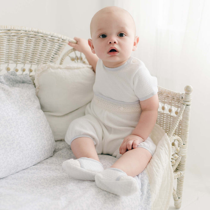 Infant dressed in the Harrison Short Sleeve Christening Romper with intricate lace trim and a white quilted newsboy cap, sitting in a classic wicker crib with delicate details.
