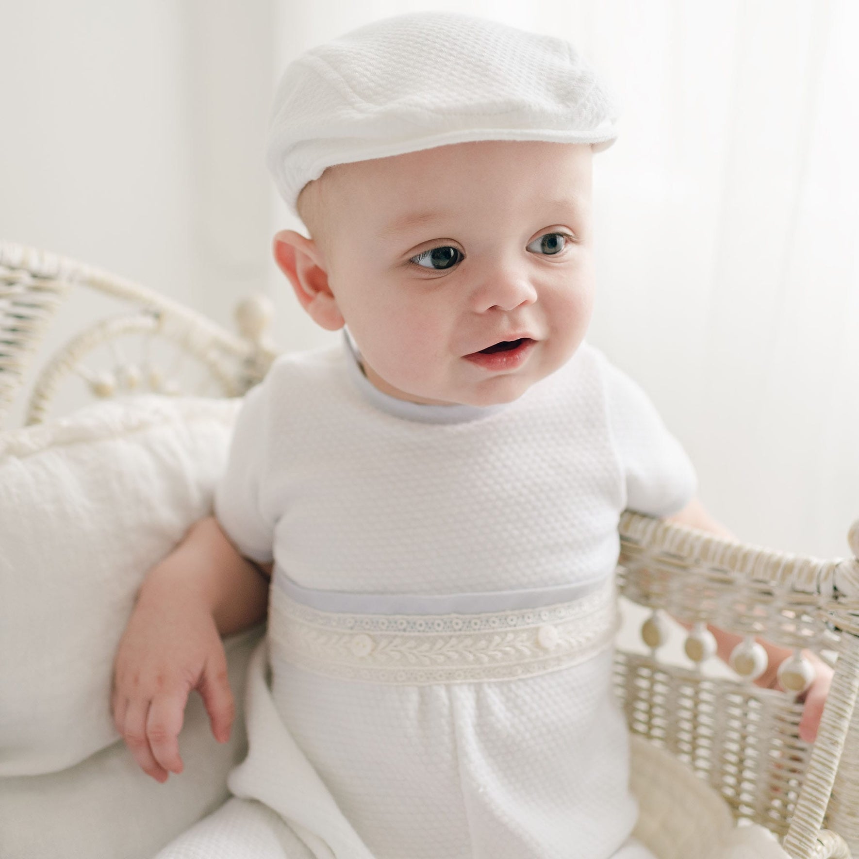 A baby boy wearing the Harrison Short Sleeve Christening Romper and cap sits in a light-colored wicker chair. The child has light skin, bright eyes, and looks to the right with a curious expression. The background features softly lit, light-colored curtains. His outfit, handmade in the USA, exudes charm and elegance.