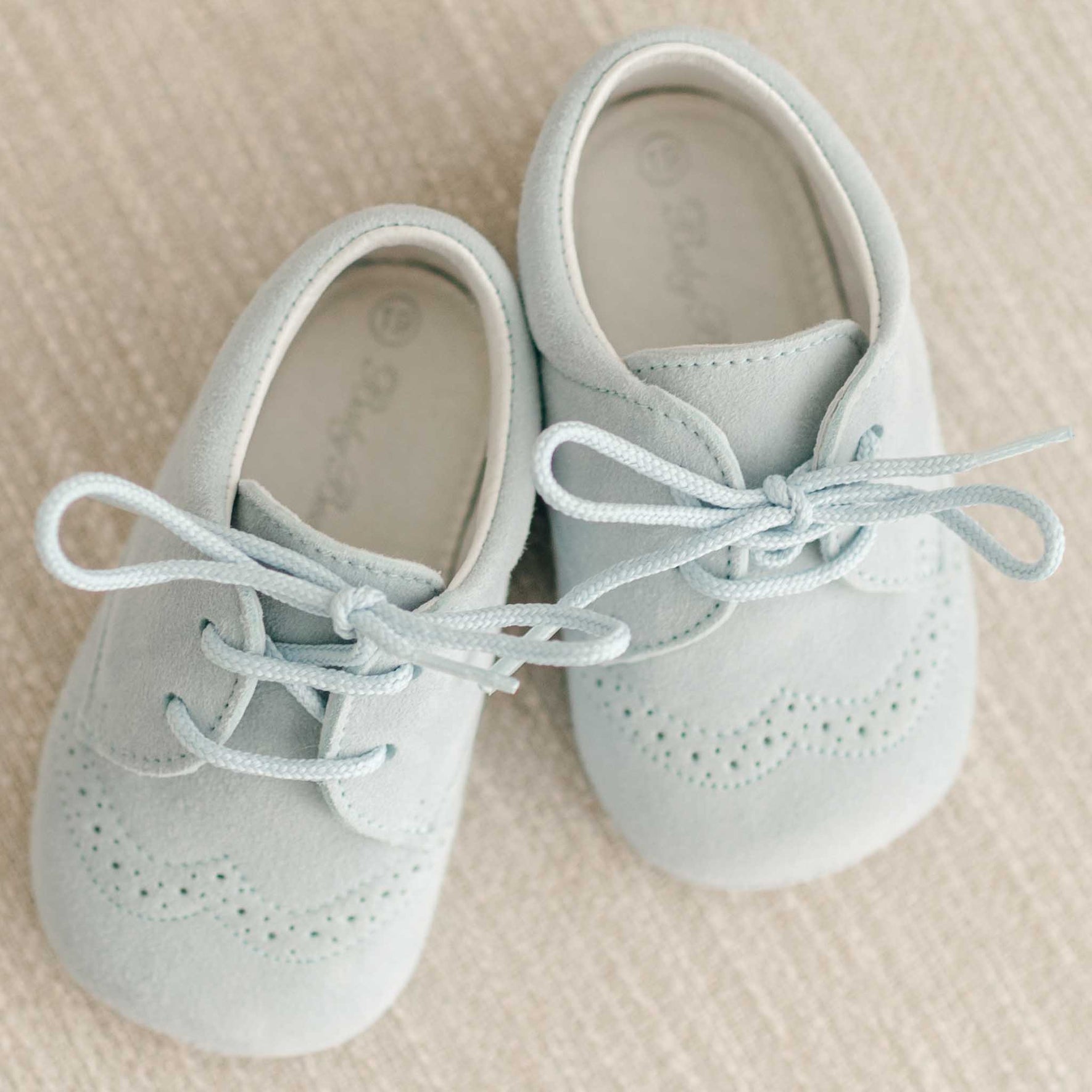 Light blue suede lace-up christening shoes for babies, elegantly presented on a soft beige backdrop.