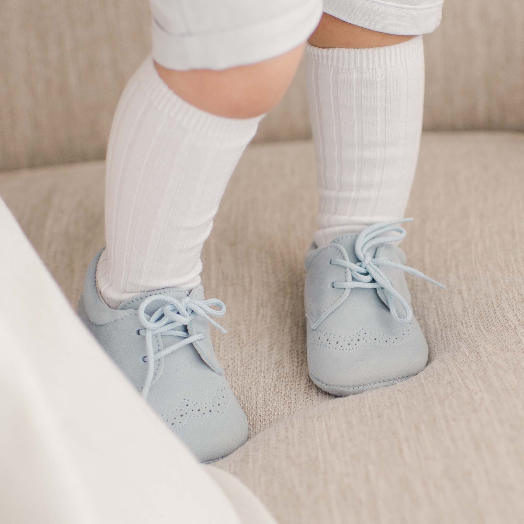 A baby standing on a chair, wearing light blue suede christening shoes paired with white ribbed socks.