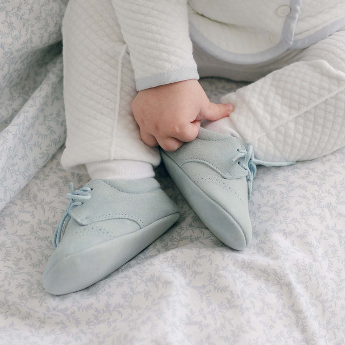 Light blue suede christening shoes with intricate perforations, worn by a baby sitting on a floral-patterned blanket.
