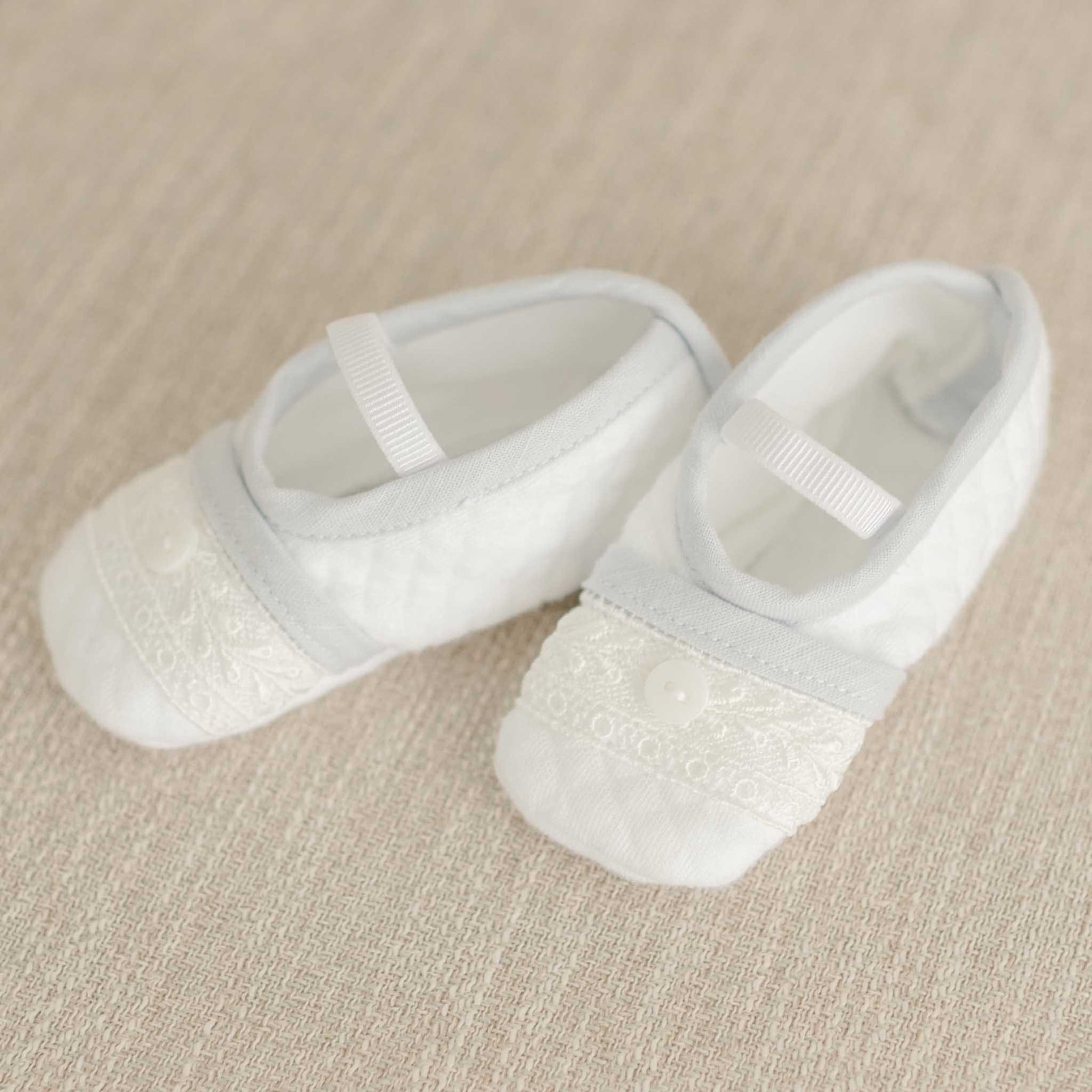 A pair of white baby shoes featuring embroidered lace detailing and small buttons rests on a beige textured surface. Ideal for complementing the Harrison Suit Accessory Bundle and completing a christening outfit, these shoes include elastic straps for a secure fit.