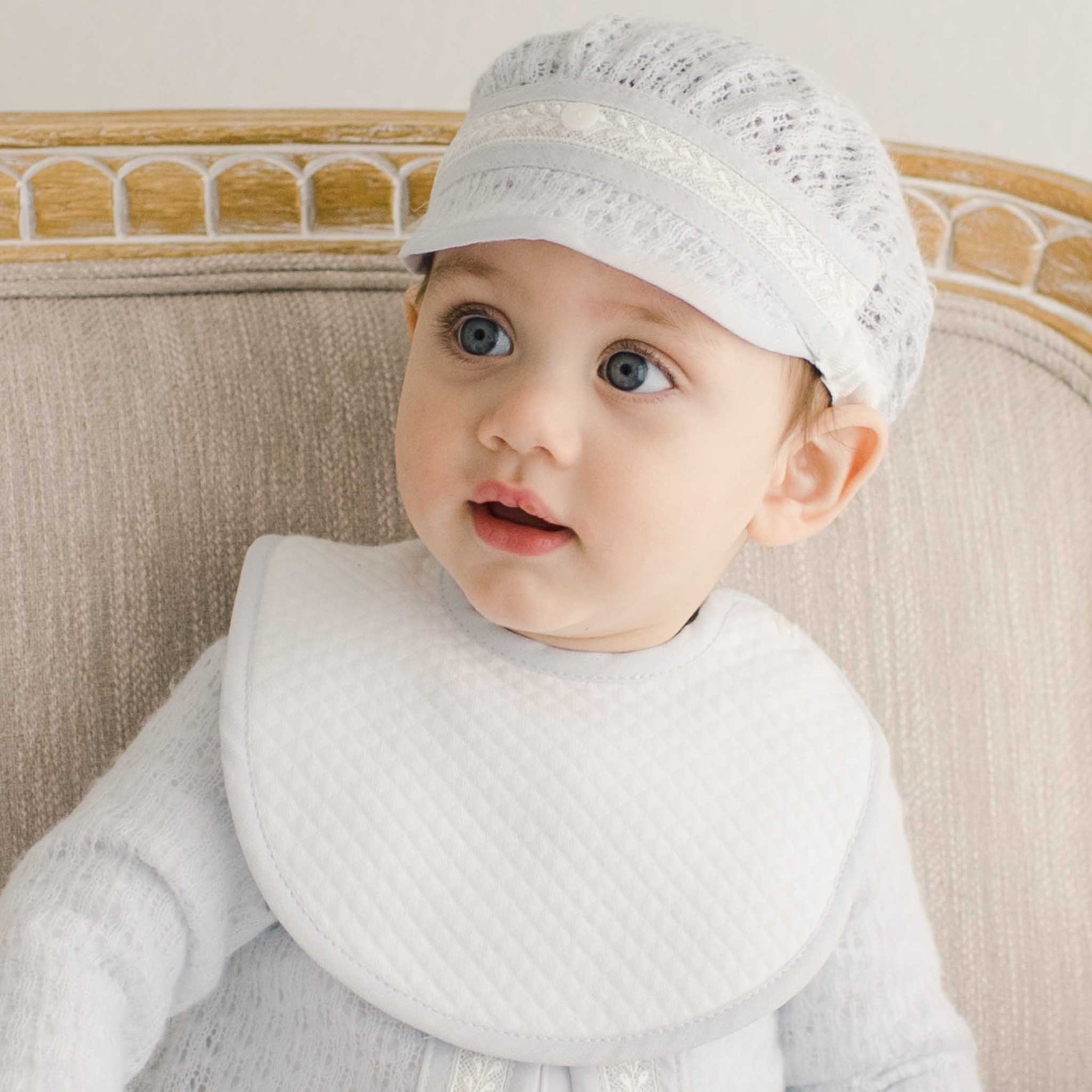 A baby in a Harrison Suit Accessory Bundle, which includes a matching cap and bib, sits on a tan upholstered chair. The baby's wide eyes and slightly parted lips look to the side, while the neutral background accentuates the image's soft tones.