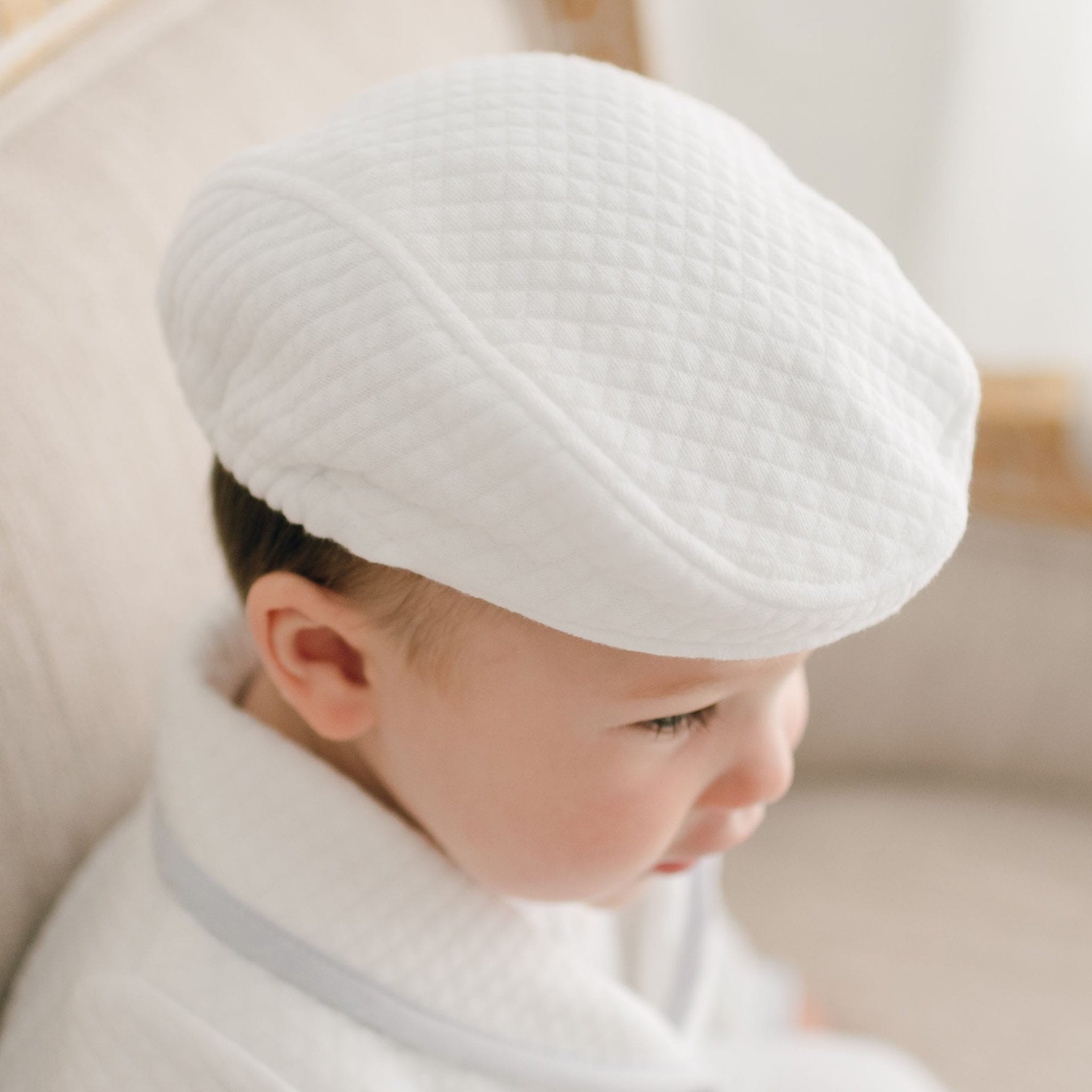 A baby dressed in the Harrison Suit Accessory Bundle, featuring a white textured cap and christening outfit with light blue trim, is seated on a light-colored upholstered chair, with the softly blurred background highlighting the baby's serene expression.