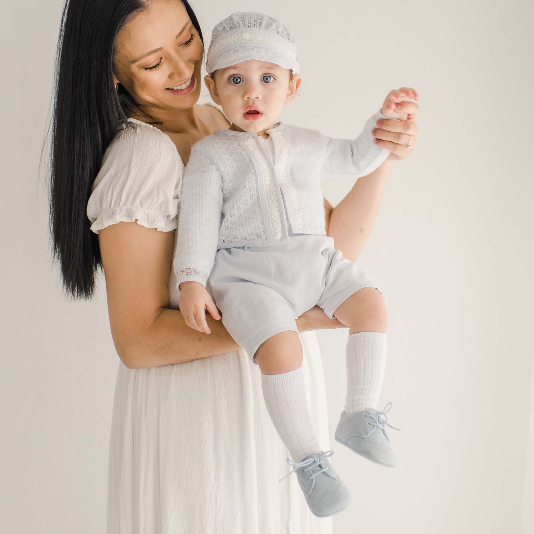 A baby dressed in a light blue knit sweater with lace accents and matching shorts, held by a woman in an airy white dress.