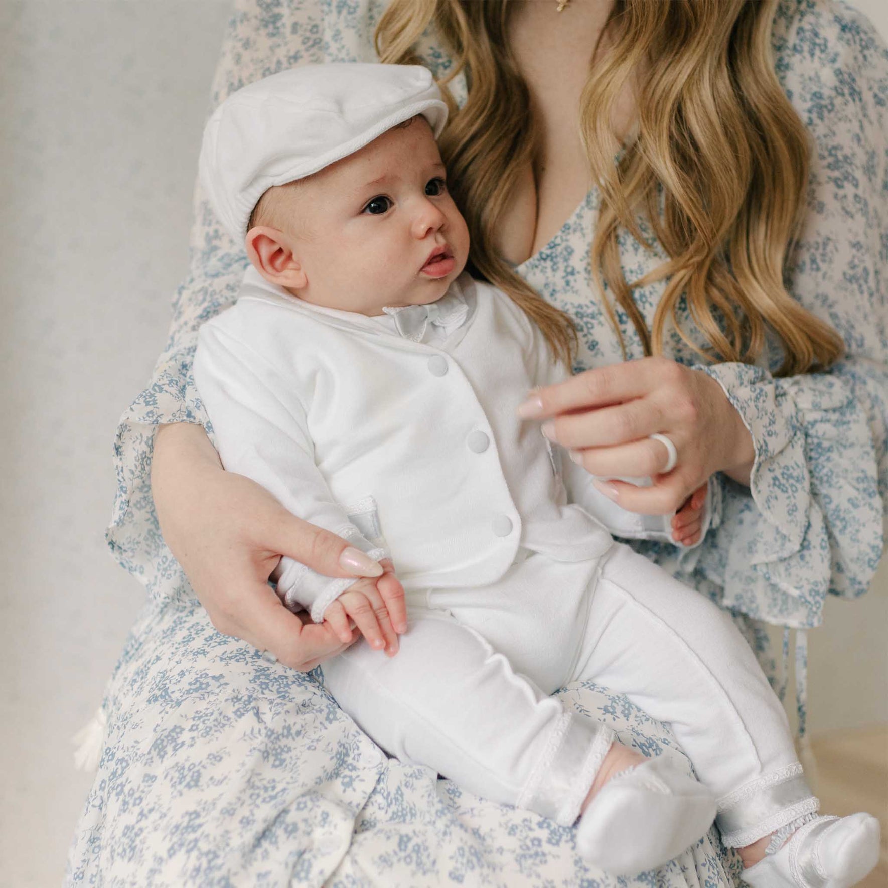 Baby in the Henry 3-Piece Suit and matching Newsboy Cap held by a woman in a blue floral dress. Set against a neutral background. The suit is crafted with white French Terry cotton and details of Venice lace and blue silk ribbon. Perfect as a baby boy baptism outfit.