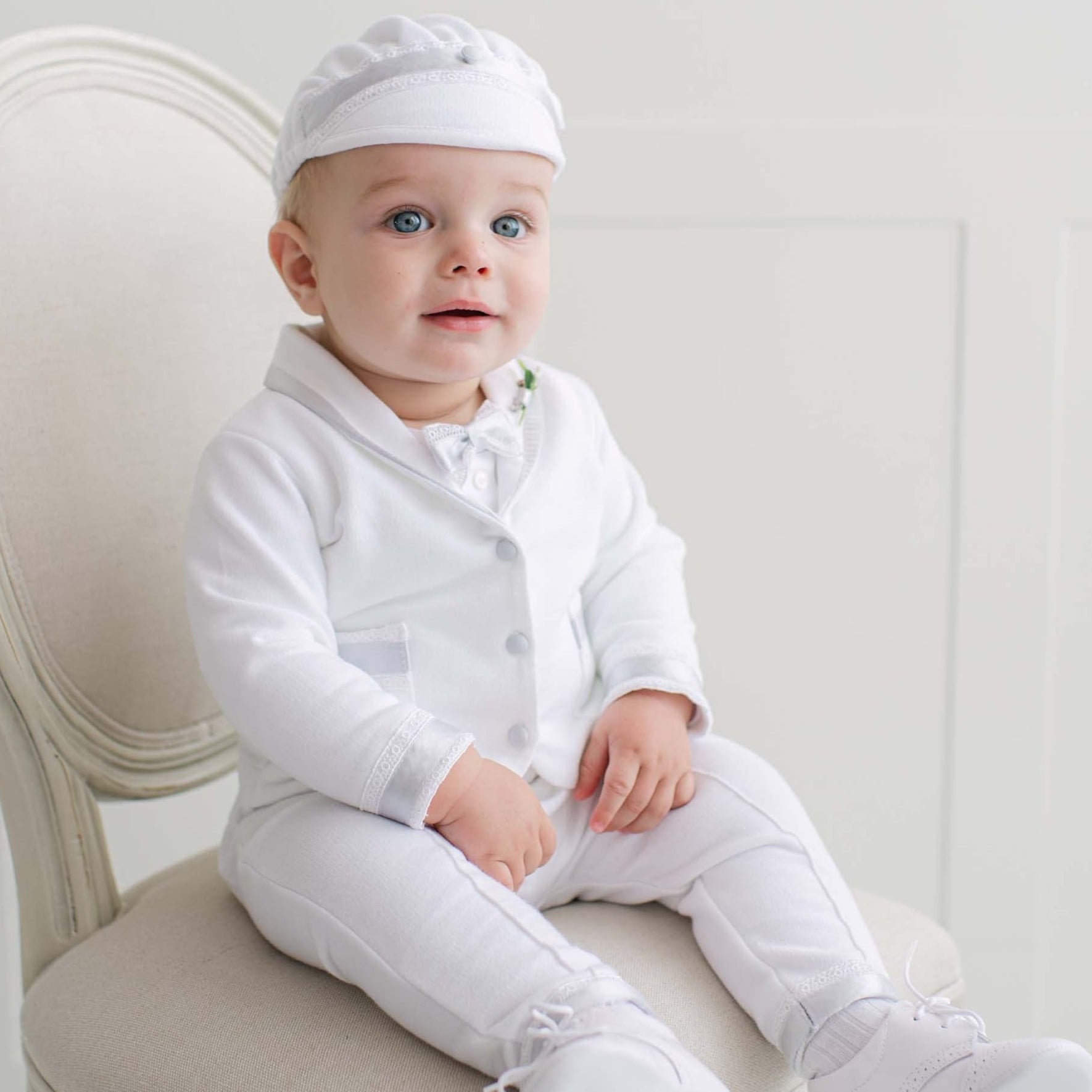 Baby in all white outfit sitting on a chair with a plain background.