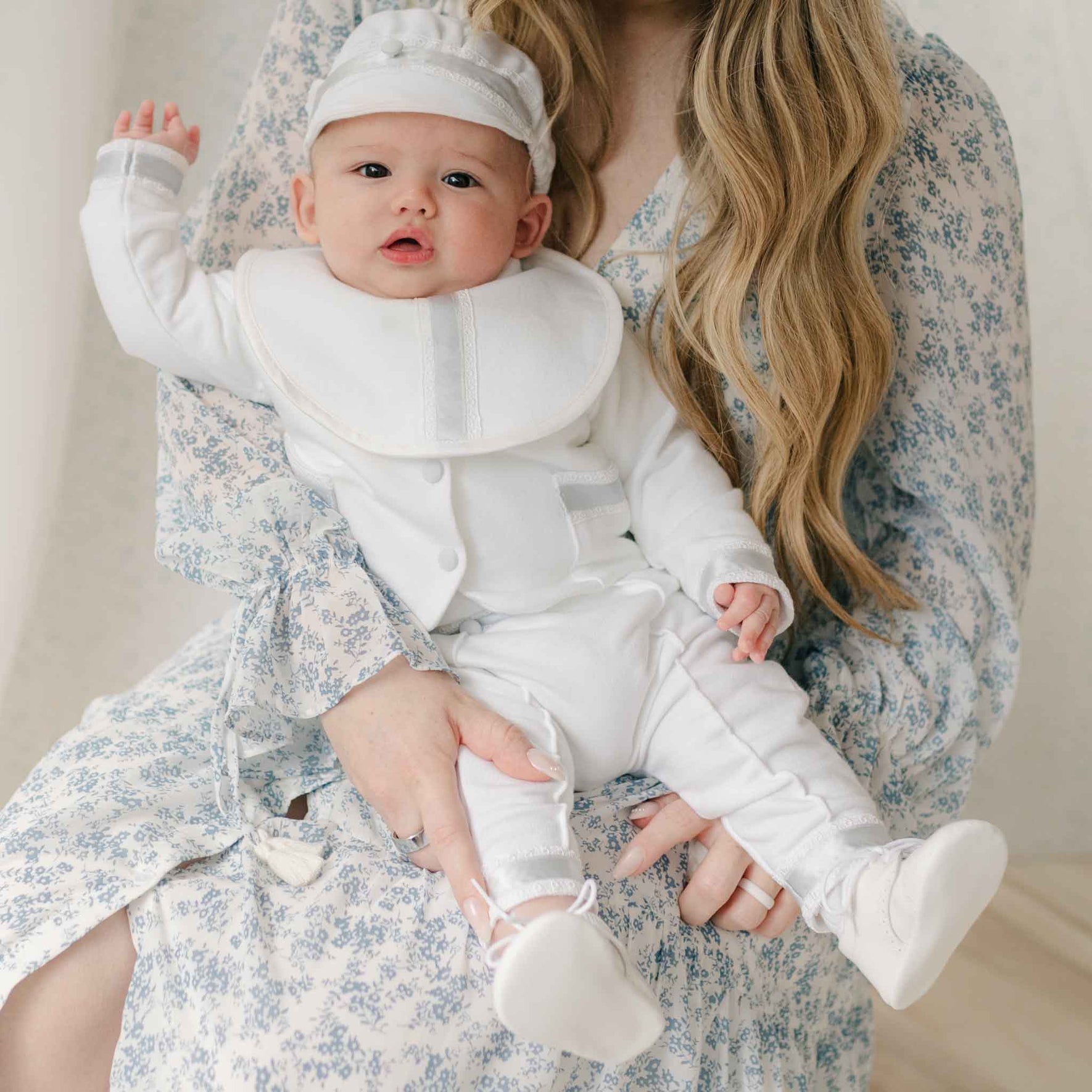 Woman in a blue floral dress holding a baby dressed in the Henry 3-Piece Suit and matching Bib. Set against a neutral background. Details on the bib and brim cap show Venice lace and silk ribbon detailing. Part of Baby Beau & Belle's Baby Boy Suit Collection.